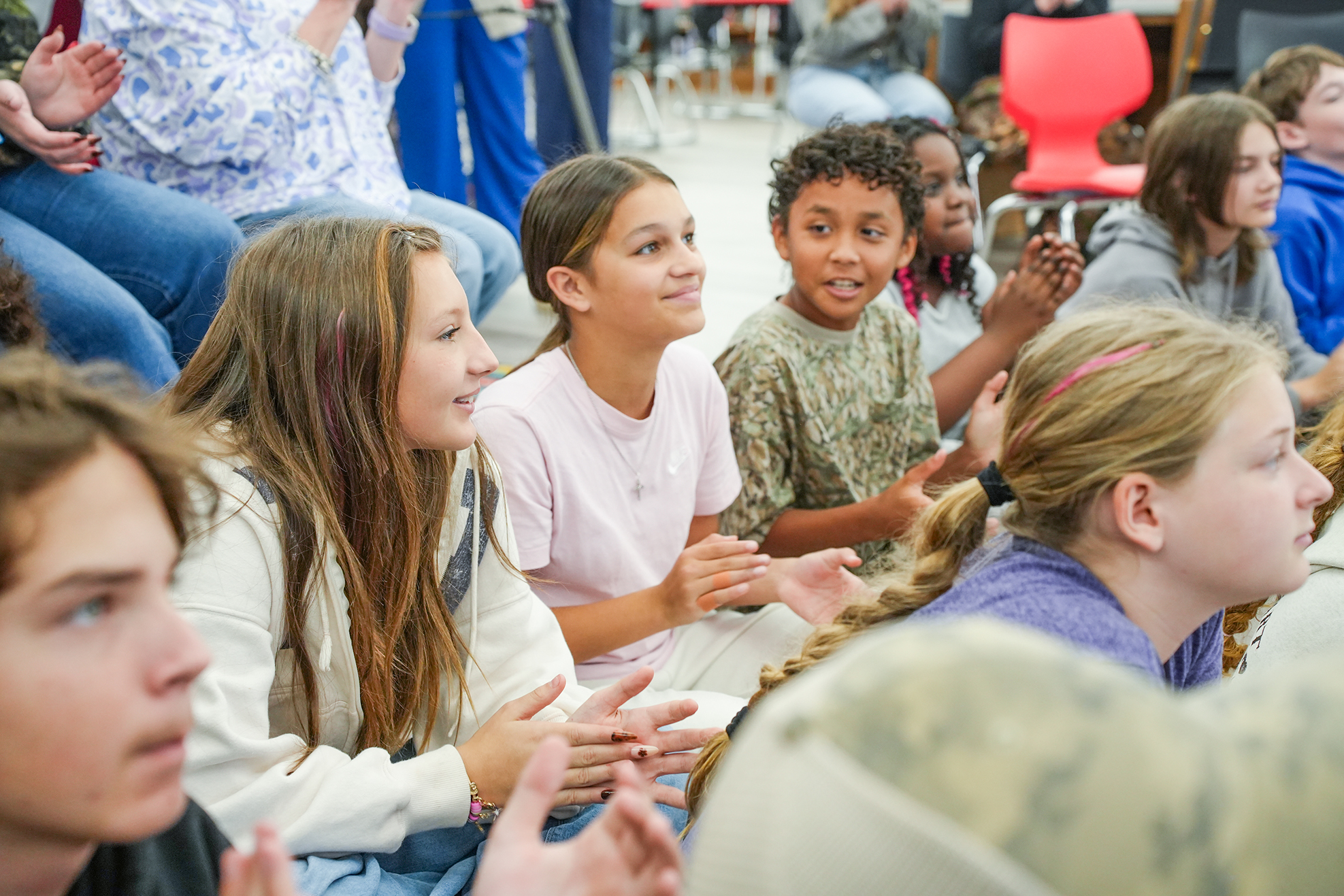 Several children seated on the floor clapping, some wearing bracelets, one wearing a camouflage shirt.
