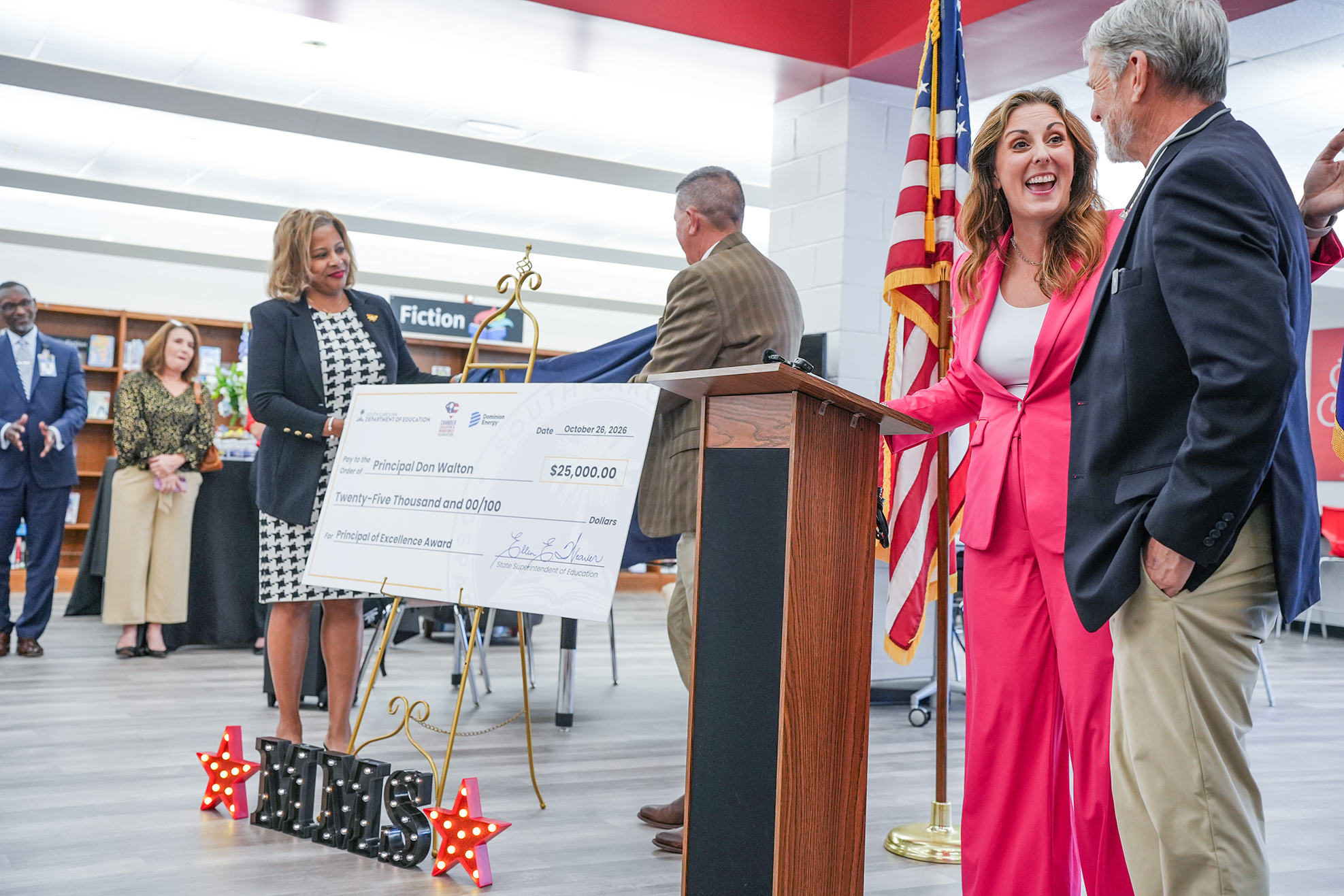 Several people stand in a room around a podium; a woman holds a large check, smiling.