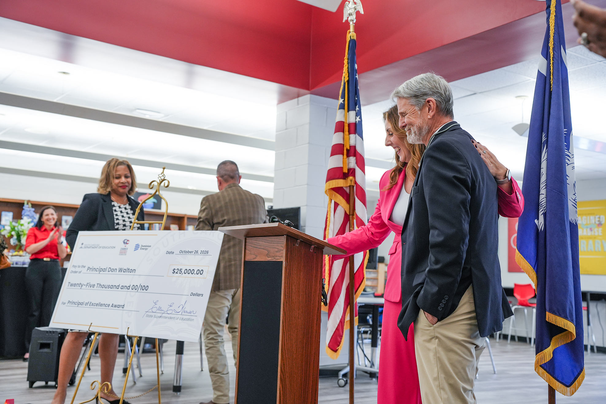 Three people stand by a podium, one holds a large check. Behind them, two flags are visible.