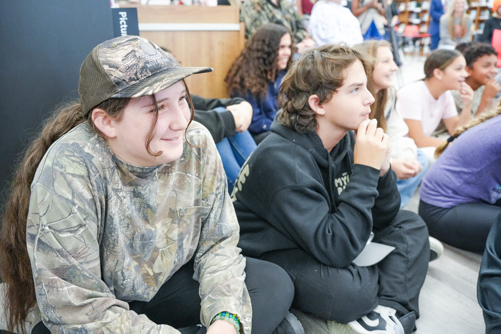 Several students sit in a room with one boy in camouflage and another in black next to him.