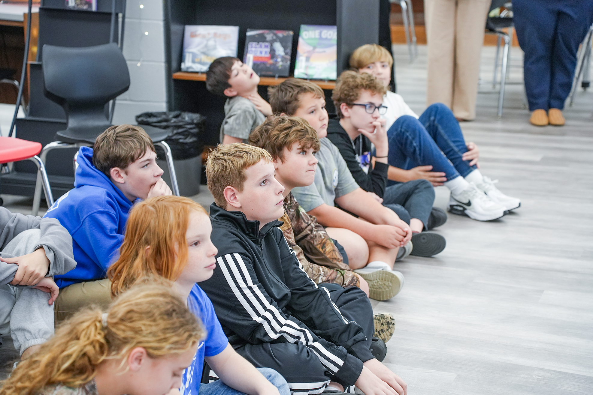 A group of students sit on the floor in a classroom, some with hands on chins, some looking upward.