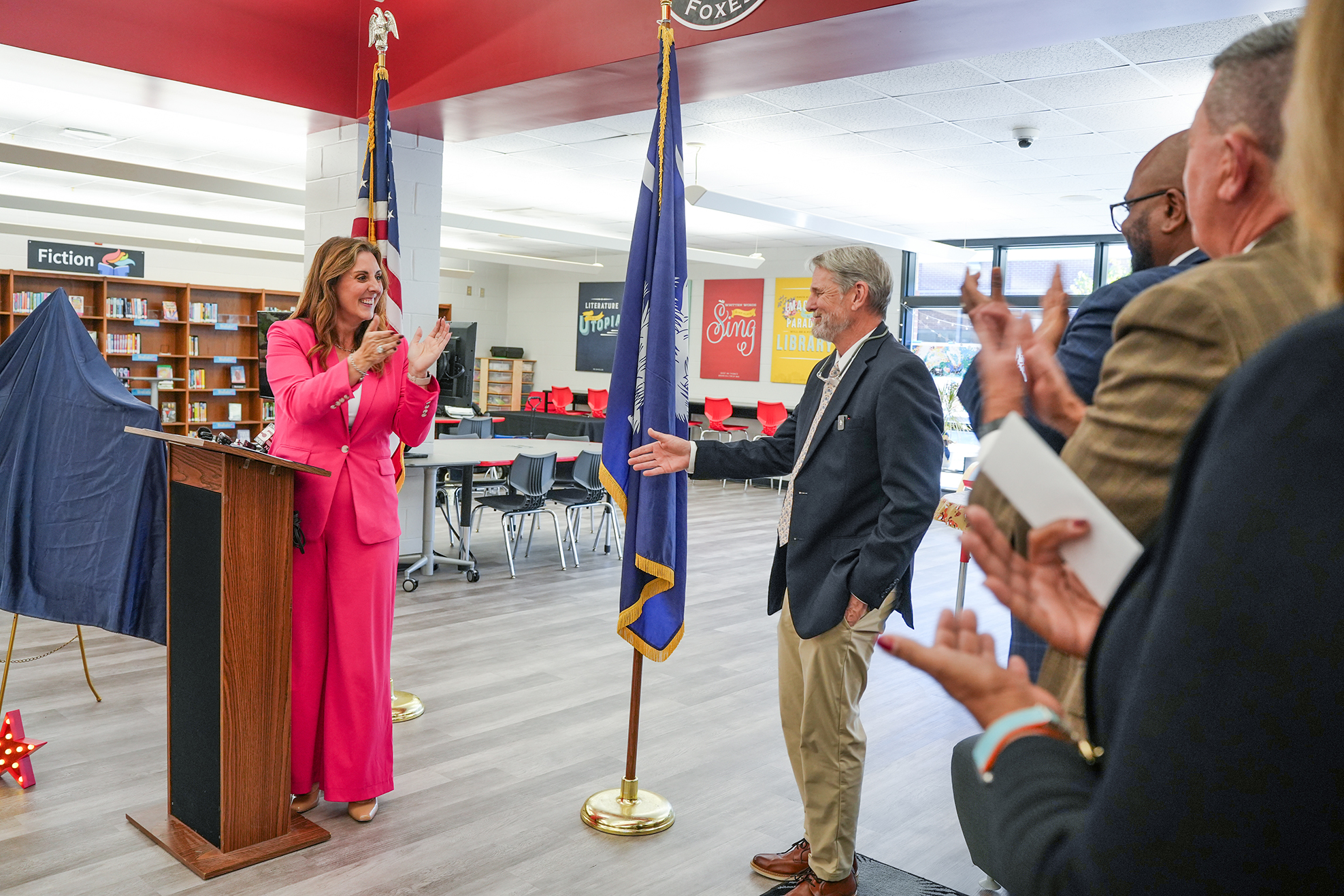 A room with a woman speaking at a podium, a man clapping next to her, and others clapping in the background.