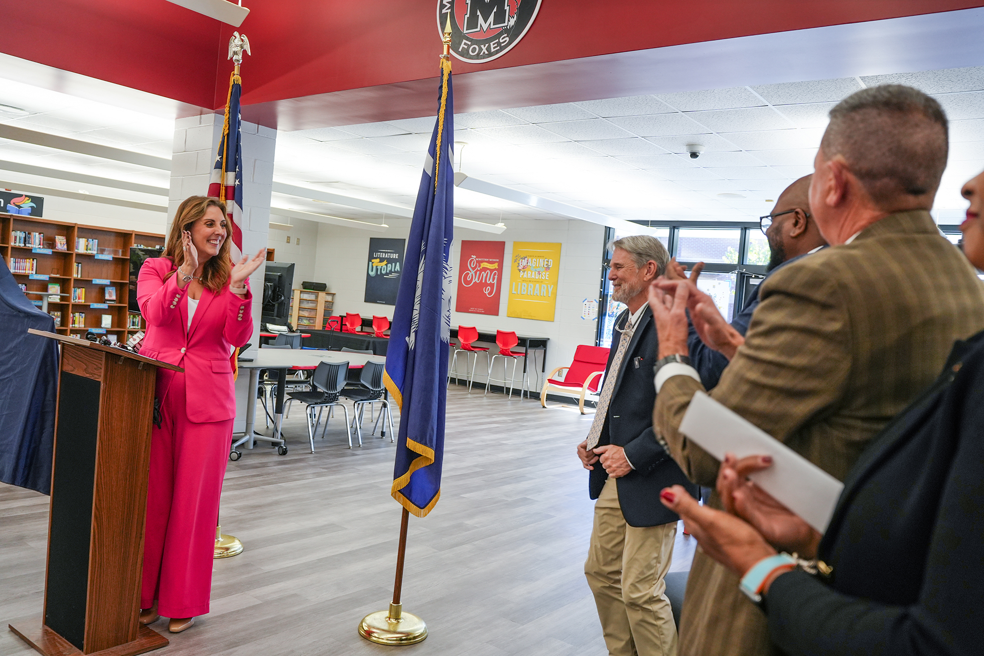 A woman in a pink suit speaks at a podium while a flagpole stands nearby. Others look on.