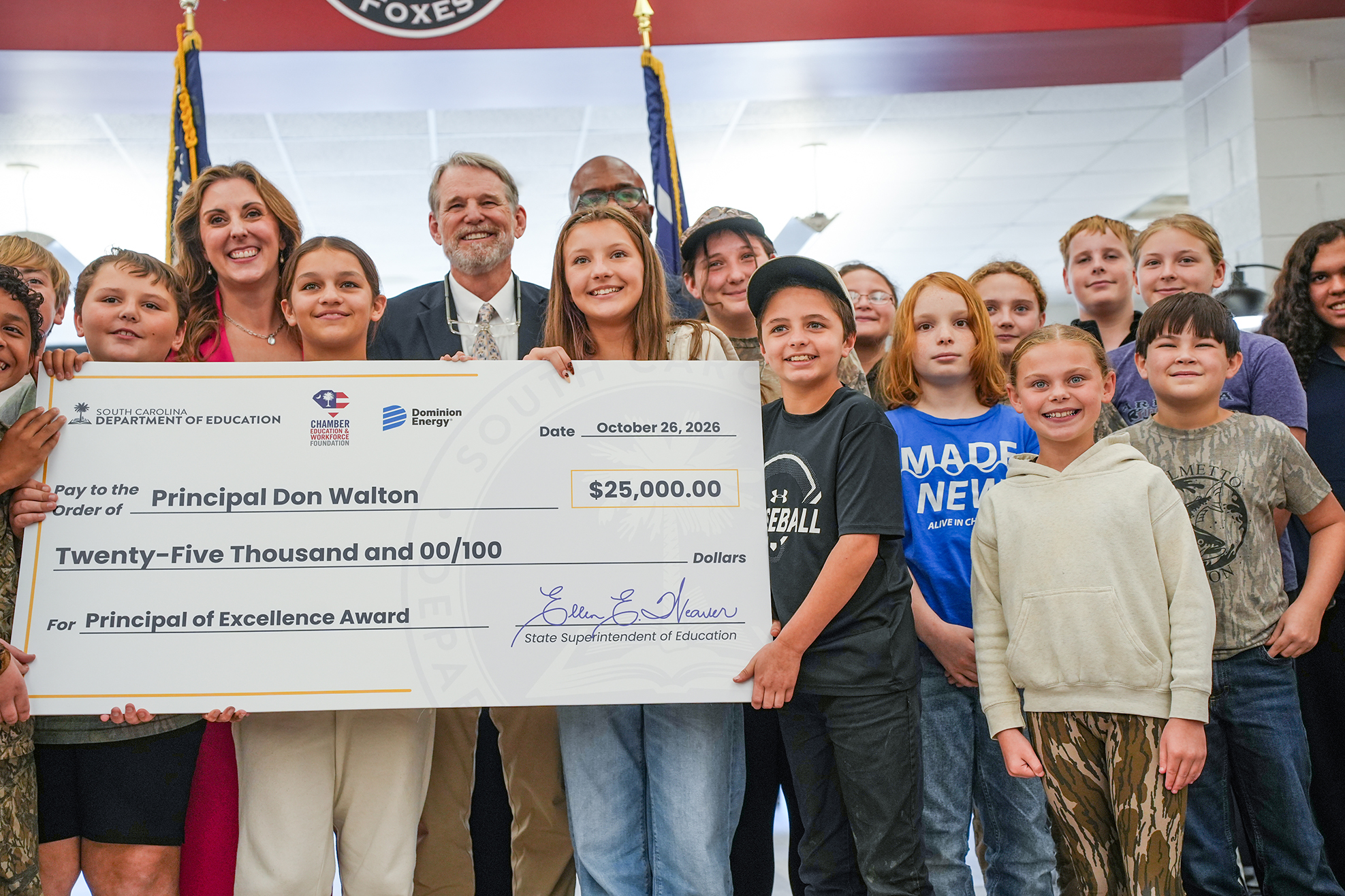 Group of adults and children posing for a photograph with a large check with signatures and a banner behind them.