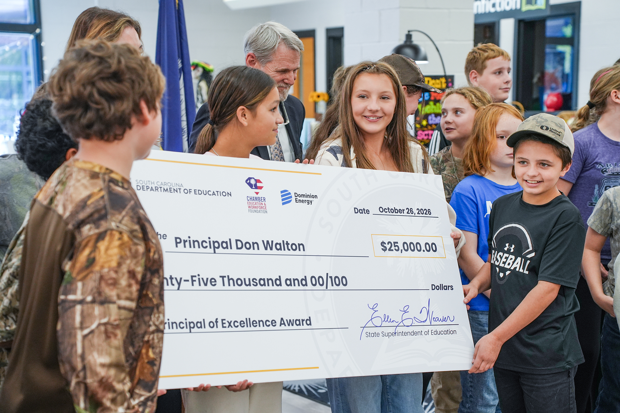 People gather indoors. A large check is displayed. A woman smiles. A child holds the check.