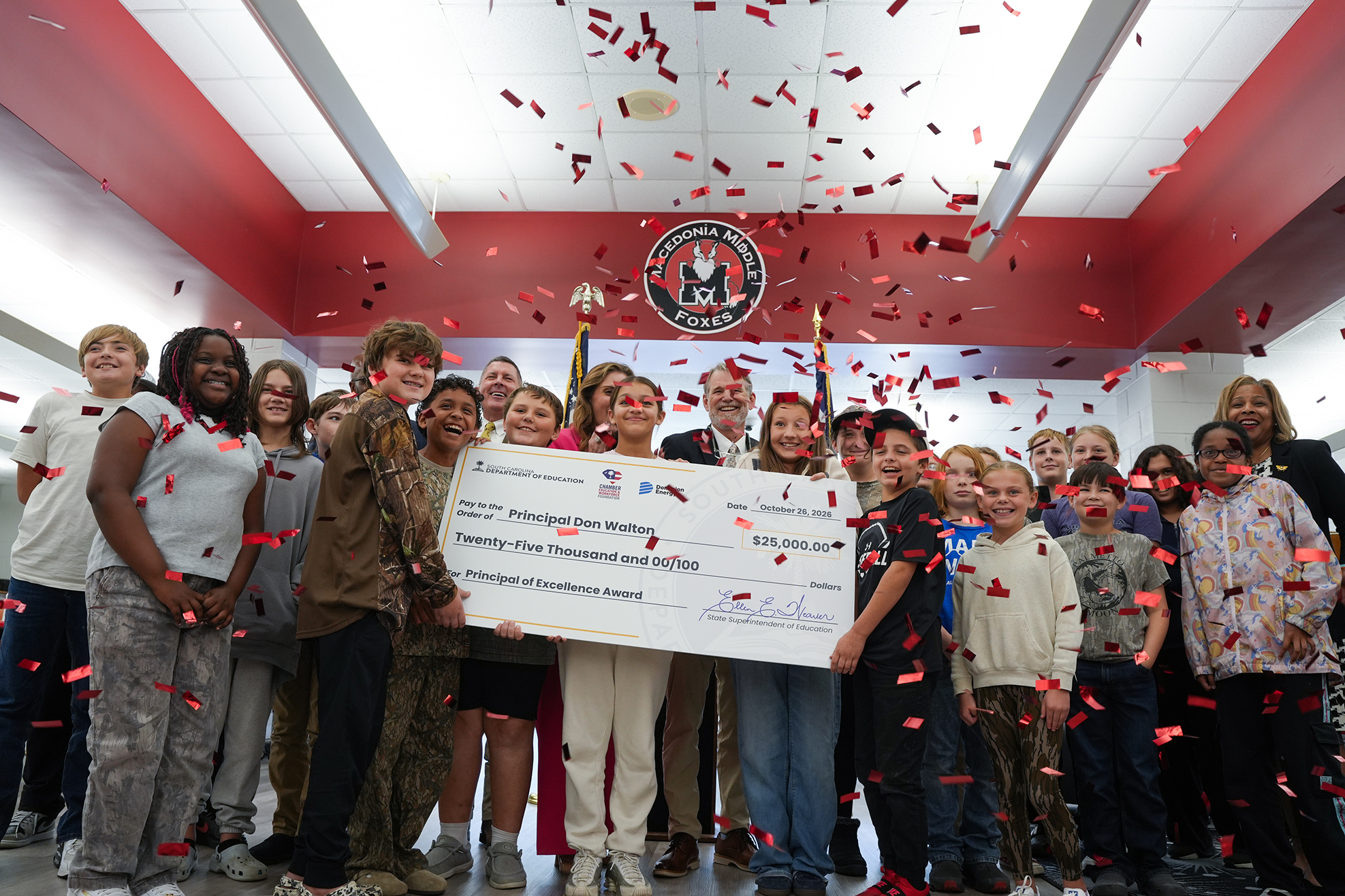A group of students, teachers, and an adult holding a large check while confetti falls.