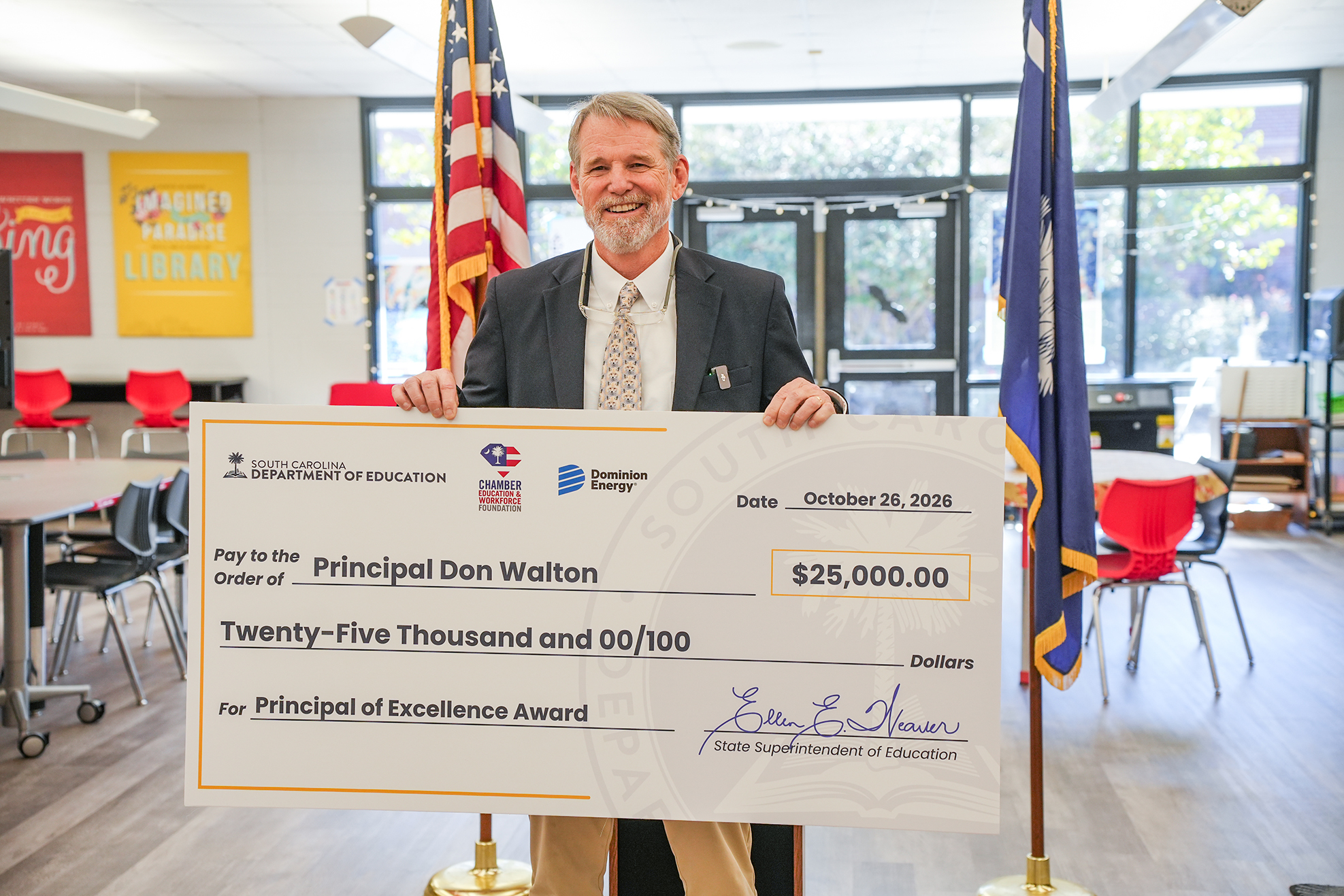 A man in a suit holds a large check. Behind him, a classroom has tables and chairs.