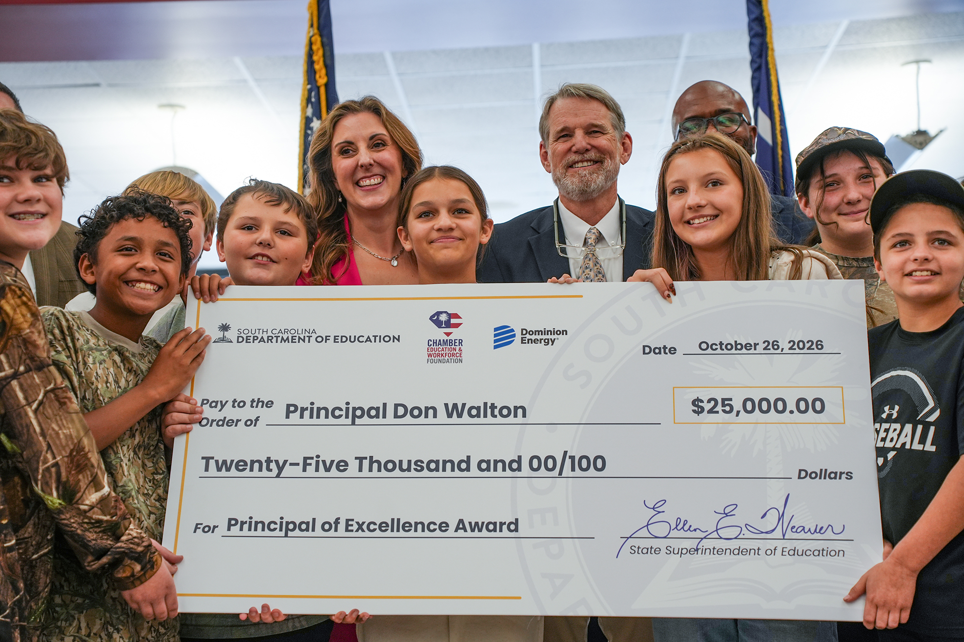 Group of people stand with large check, some wearing camouflage clothing. Check reads "$25,000.00" and "Principal of Excellence Award."
