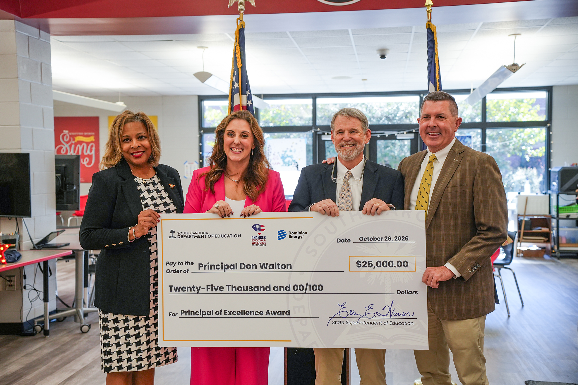 Four people hold a large check. Two women in pink and black jackets, a man in a light blue suit, and a man in a brown suit.