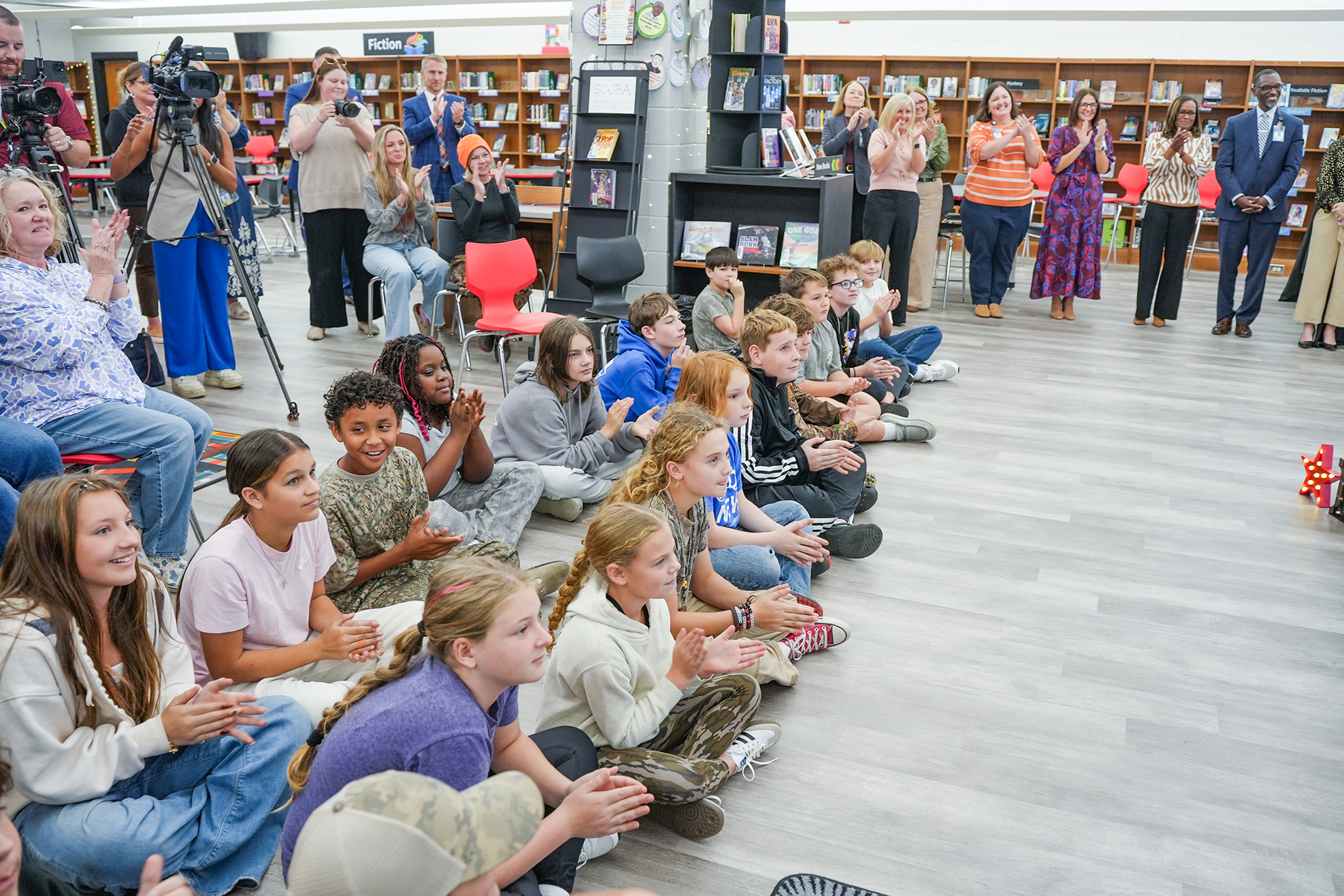 A library room with a group of students seated in front of an audience. Some adults stand behind the students, and some are seated.