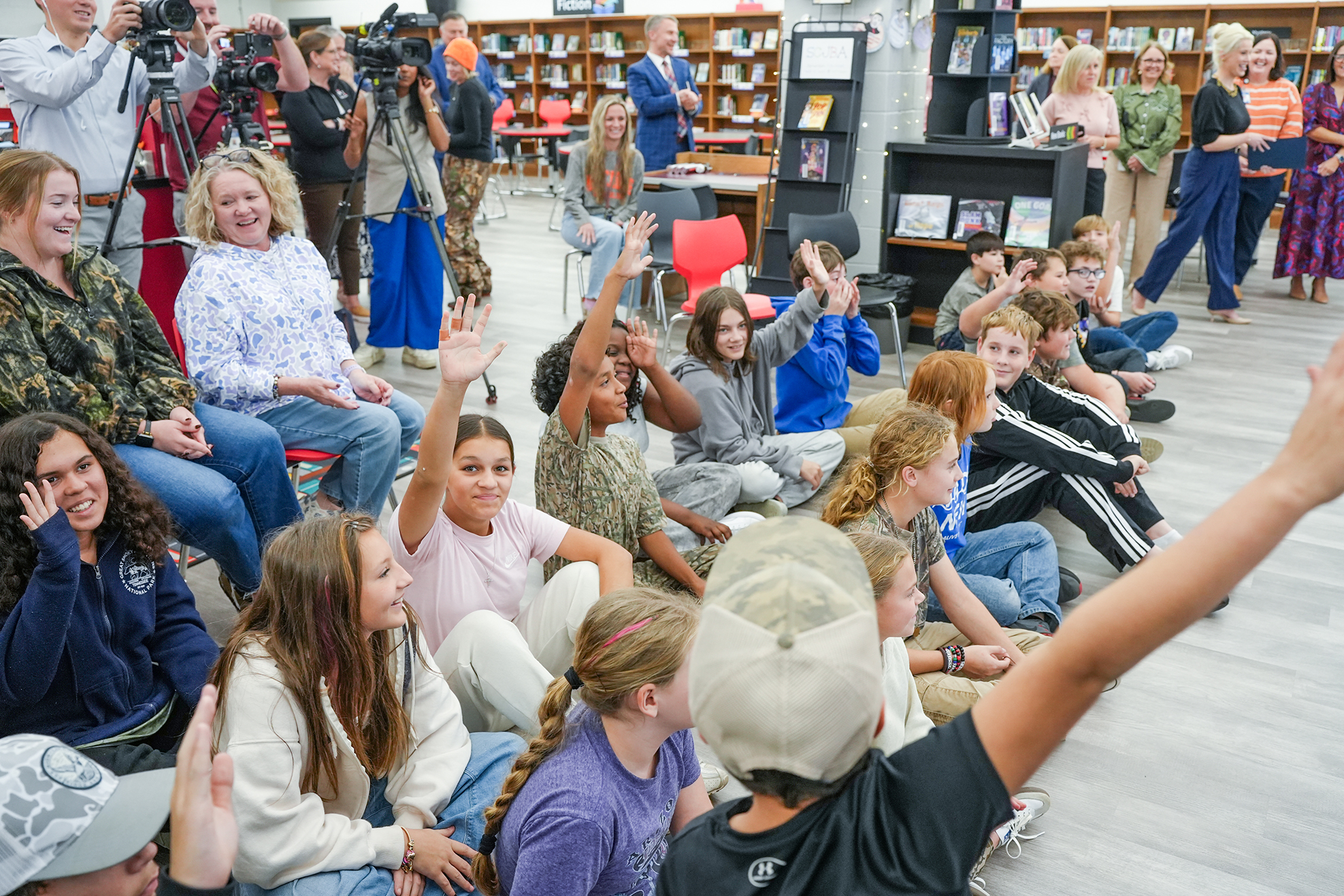 Children sit on the floor in a school library, raising their hands while adults stand and sit nearby.