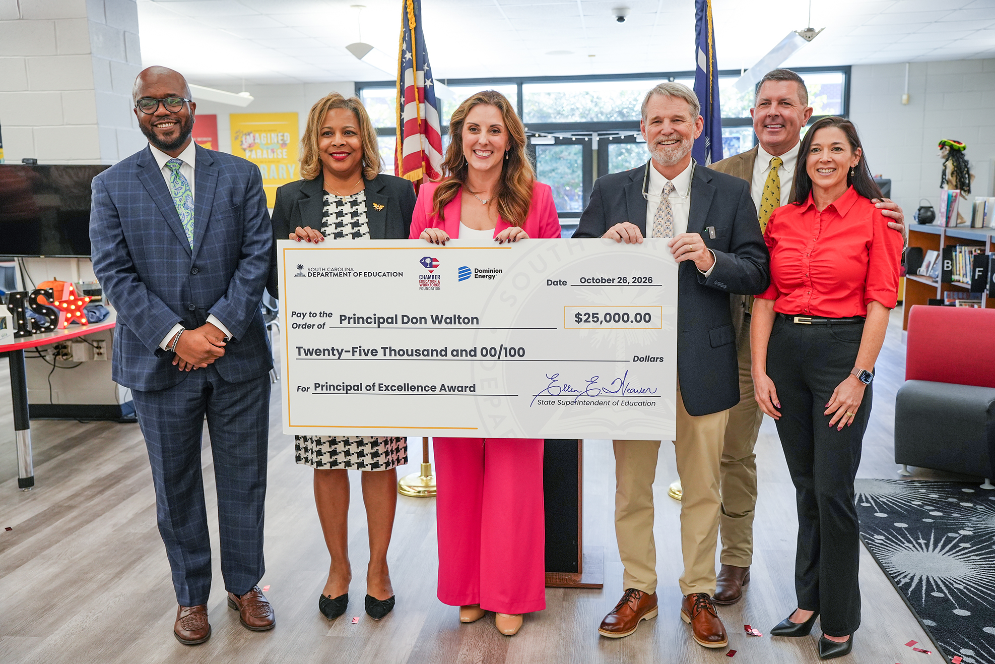 Group of adults standing, holding a large check, smiling. Indoors, in a classroom.