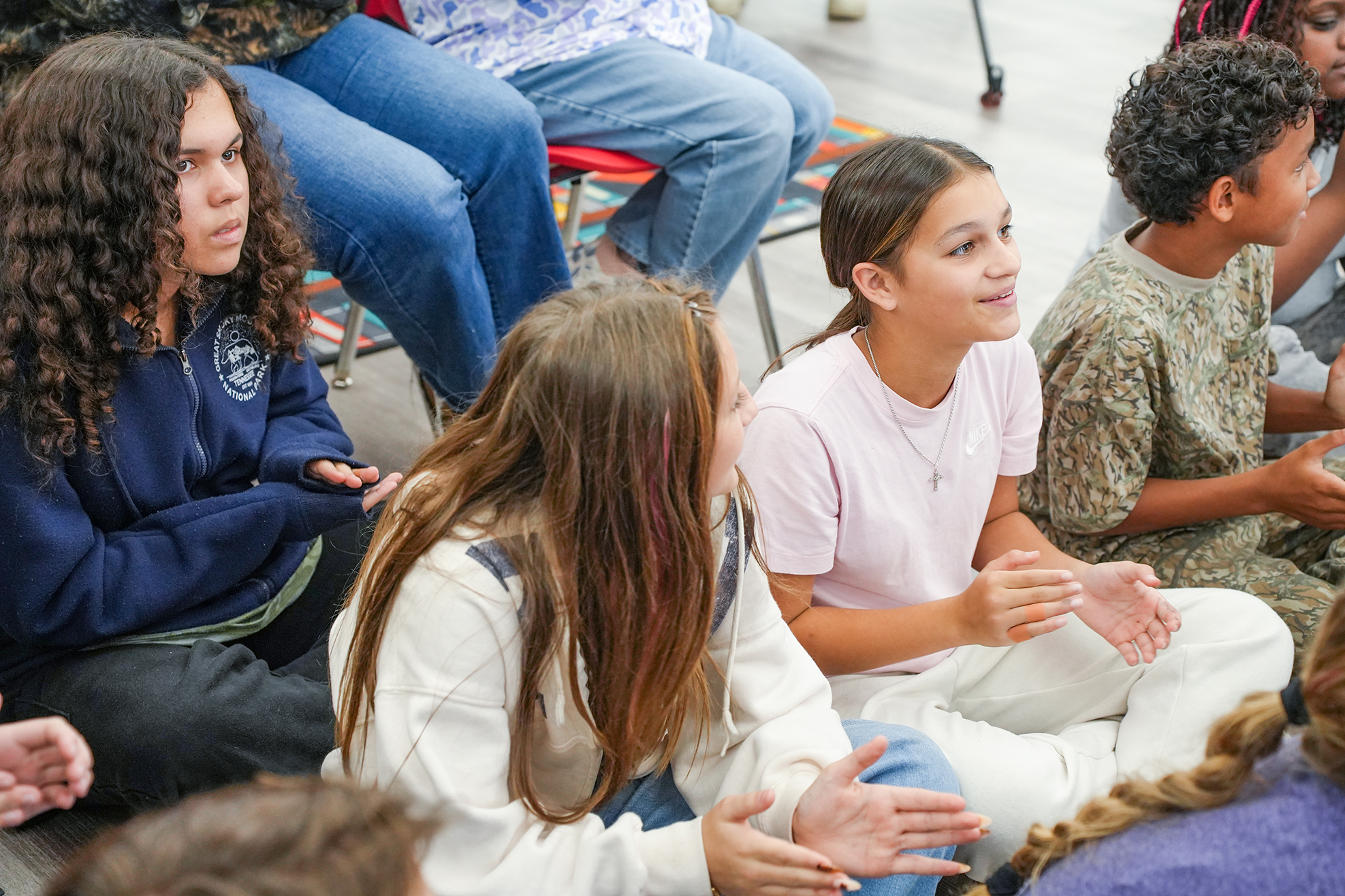 Group of students sitting in circle, discussing with raised hands, on a white floor.