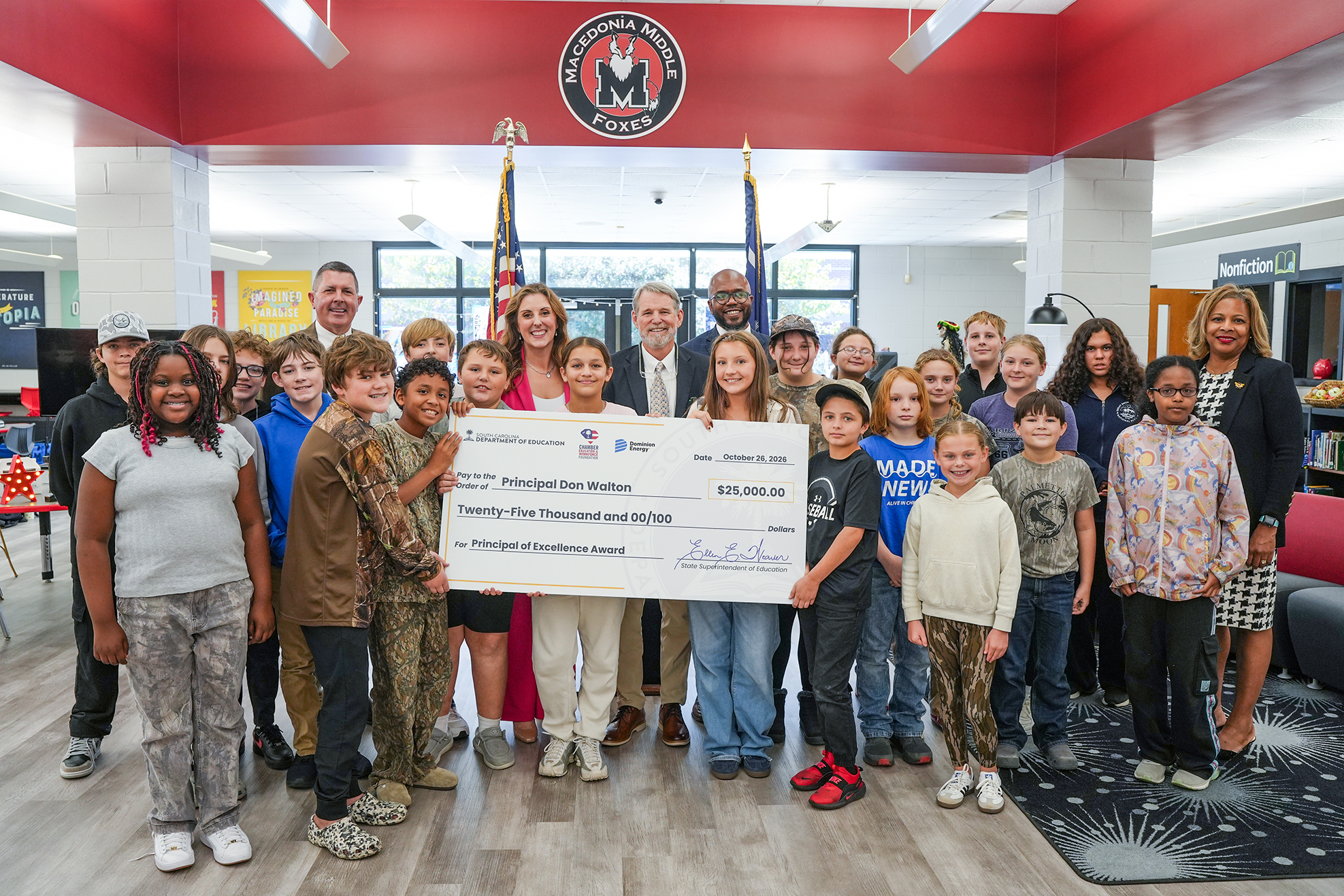 A group of students, adults, and a principal stand together, holding a large check over their heads.
