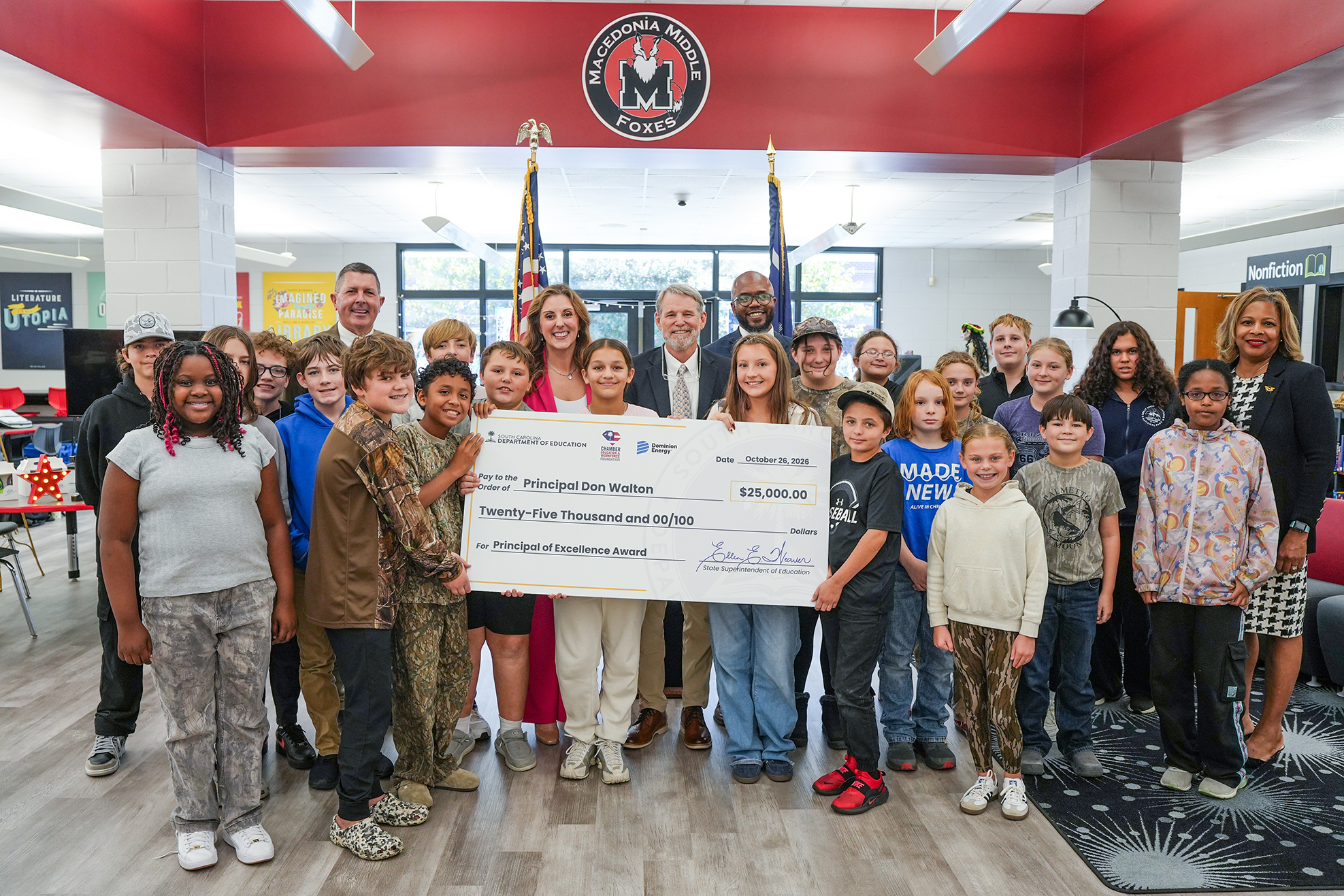 Group of adults and children posing with a large check in a room with red walls and flags.
