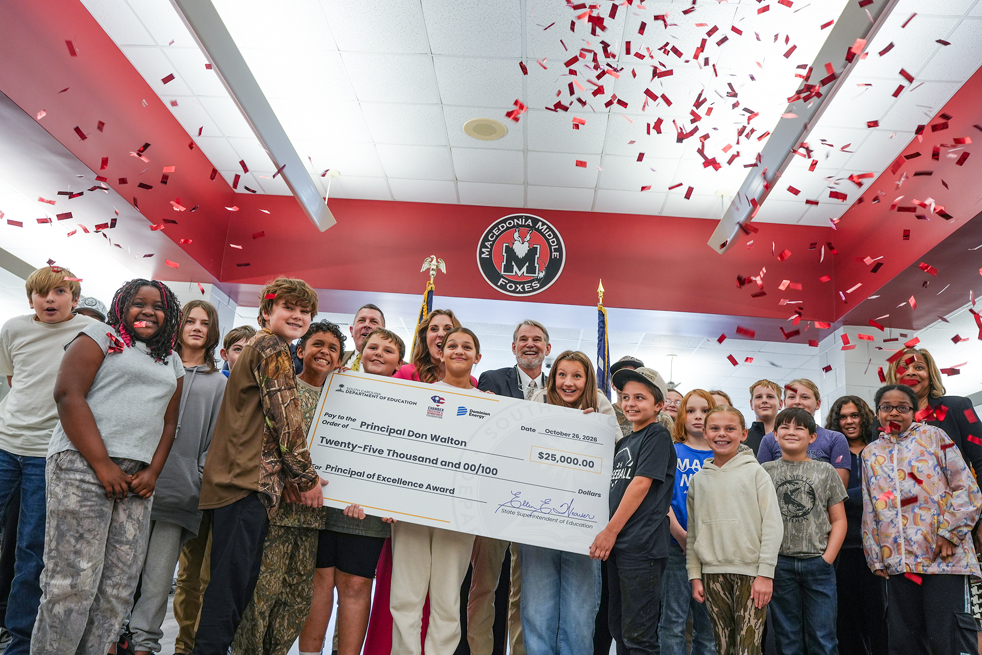 A group of children, adults, and a man holding a large check with a crowd in a room.