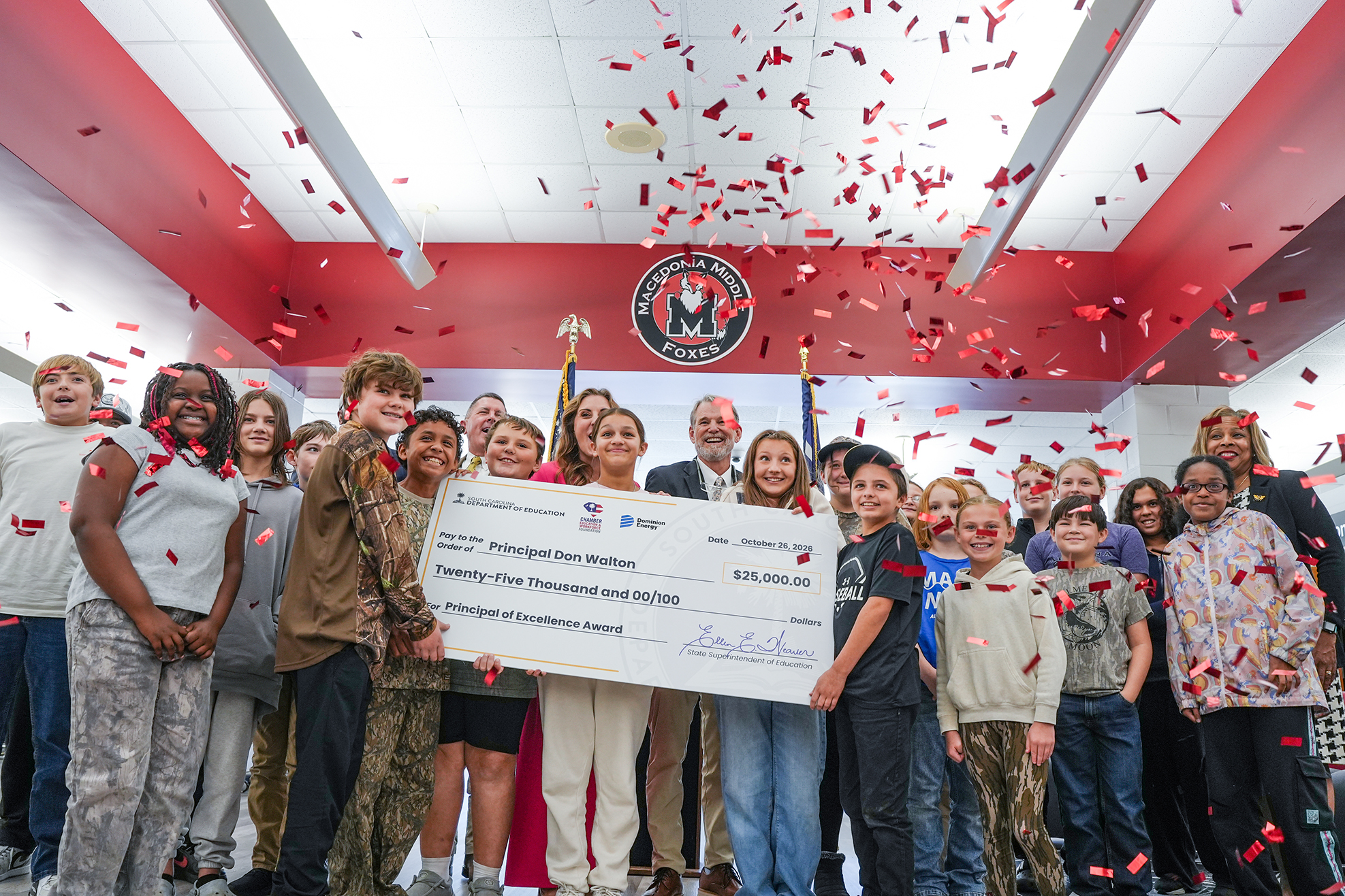 Indoor group photo; people holding large check; red confetti falling; some hold flags; bright red and white room with flag.