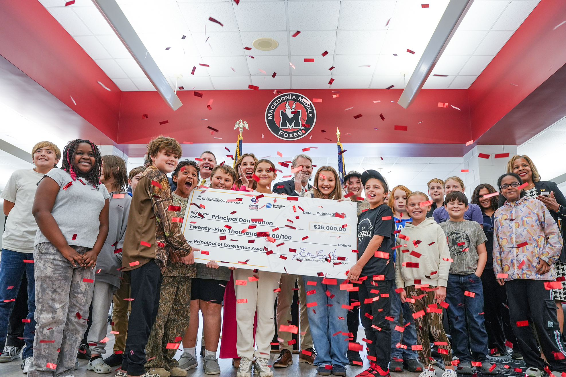 Several adults and children holding a large check with confetti falling around them in a decorated room.