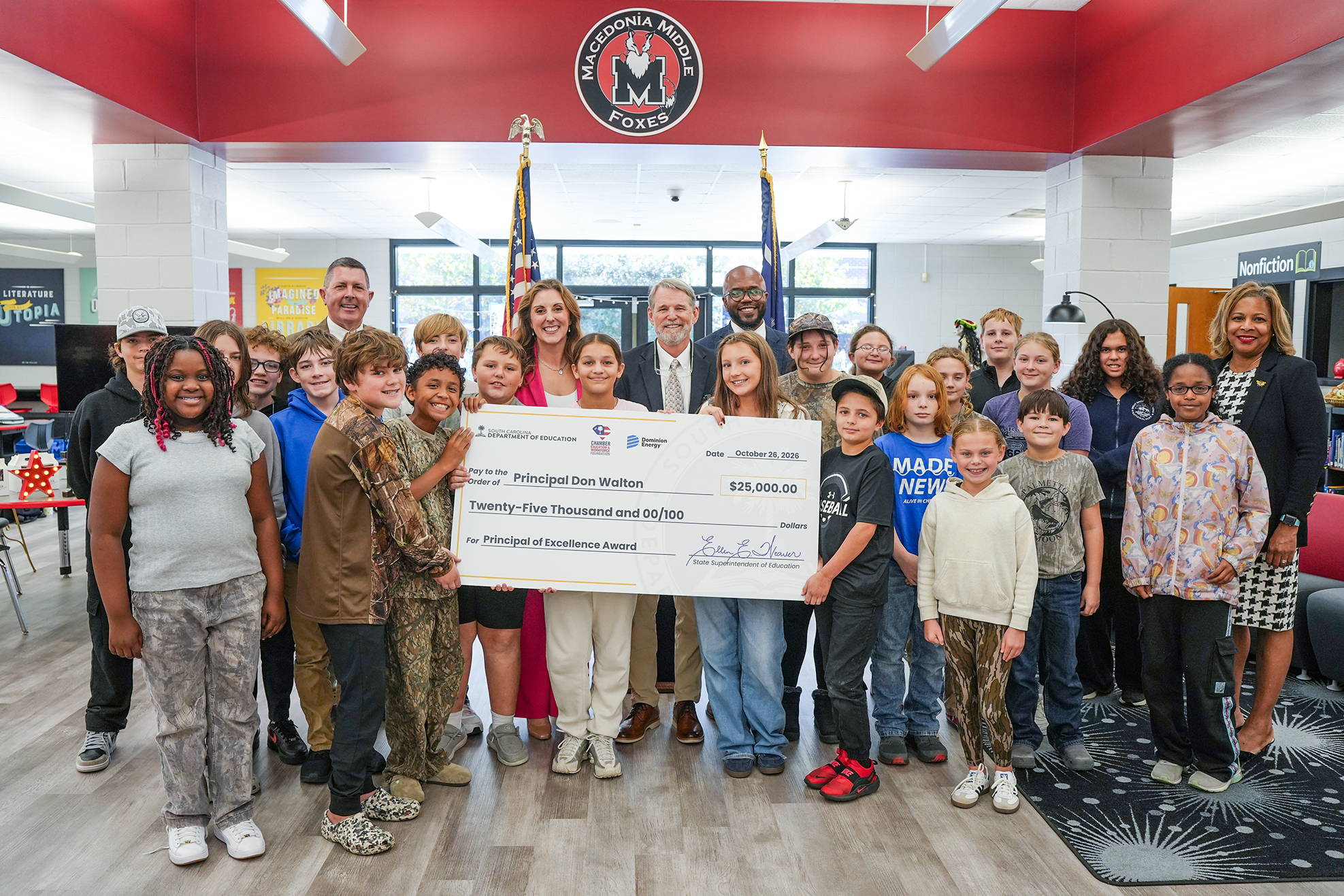 Several children and adults stand in a room, holding a large check. Behind them, two flags are displayed.