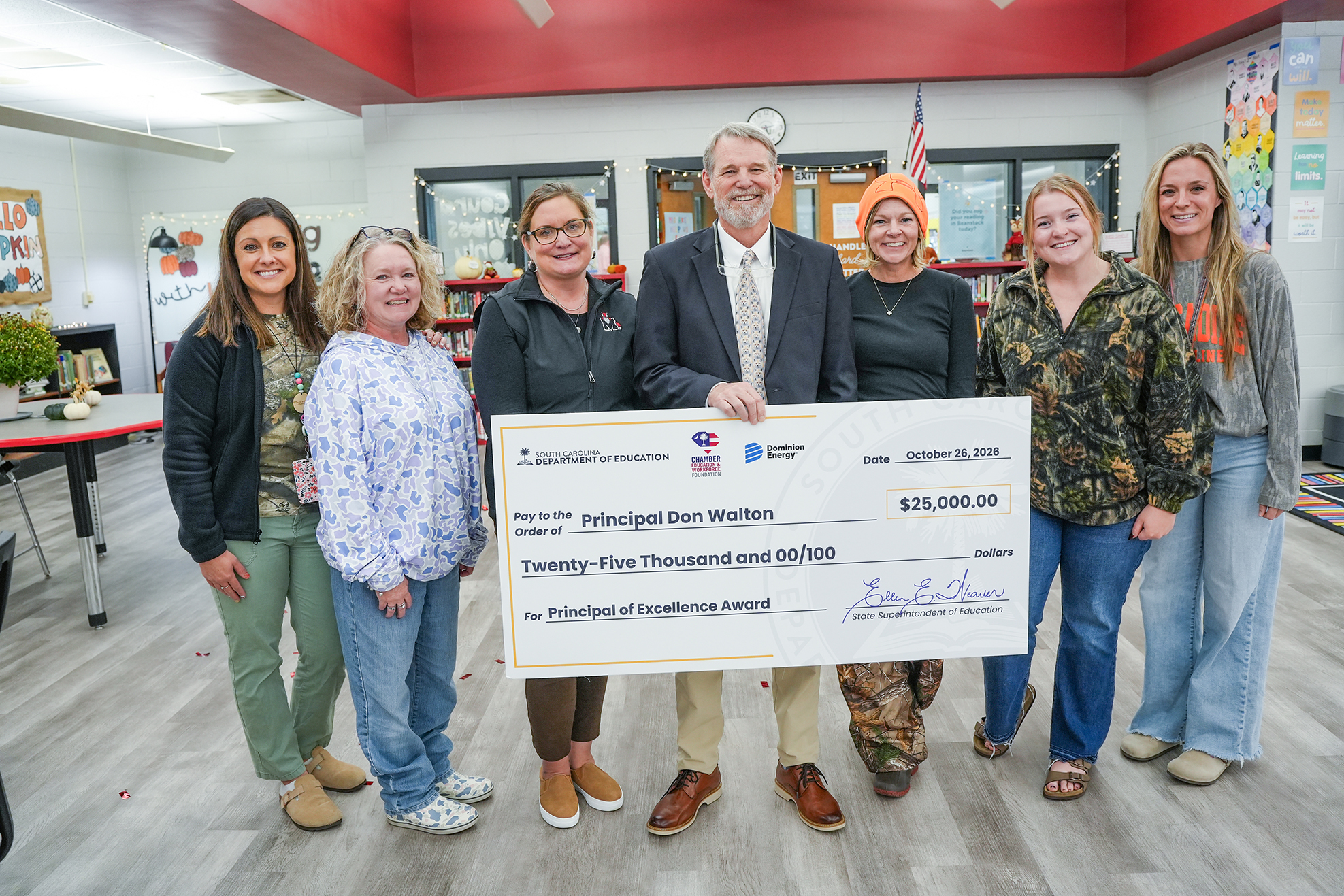 A group of people holding a large check, standing in a classroom with white walls and a red ceiling.
