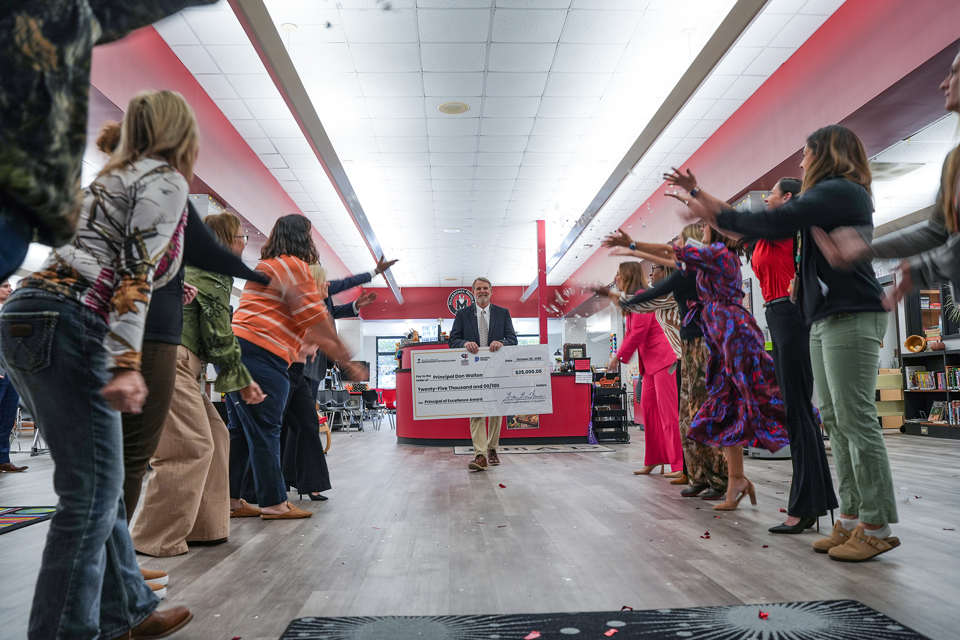 People form a circle around a man holding a large check in a library. Some have hands raised.
