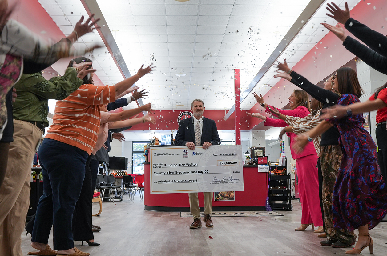 A man in formal wear holds a large check, surrounded by several people raising their hands.