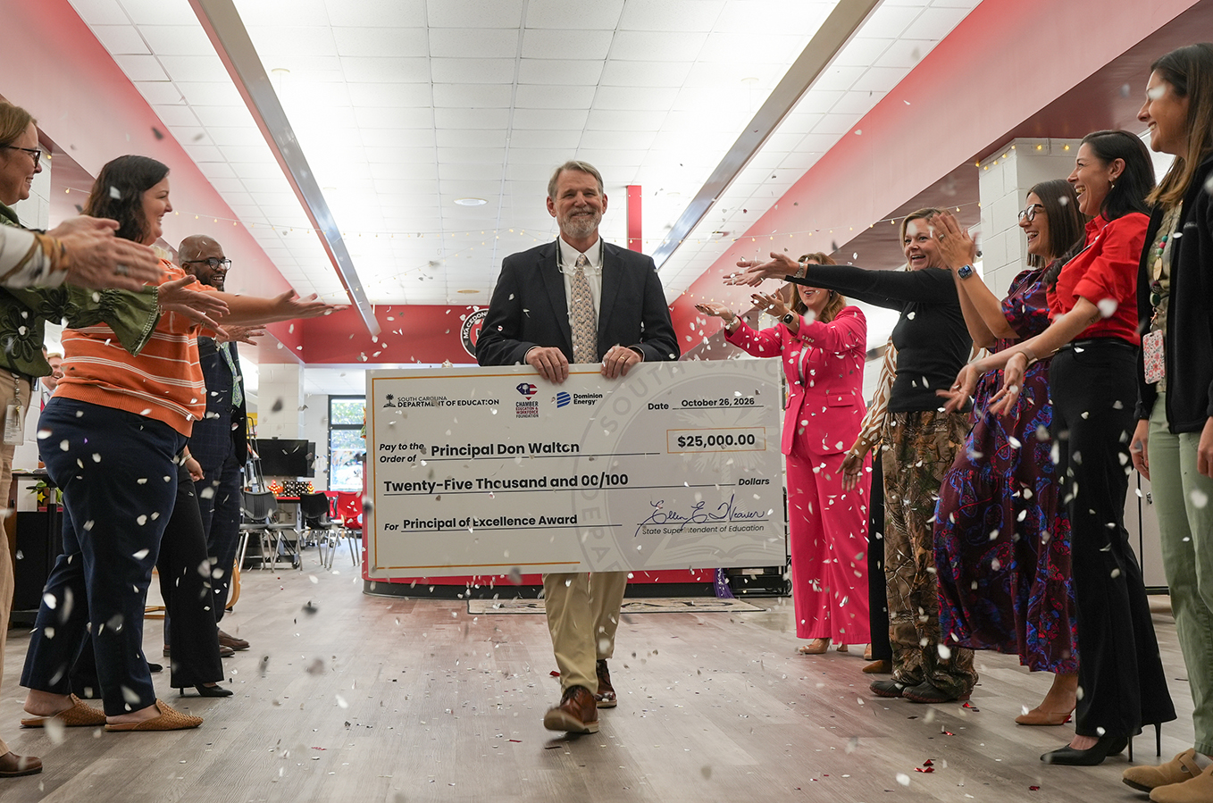 People celebrate with confetti as a man holds a large check in a room with wooden floors.