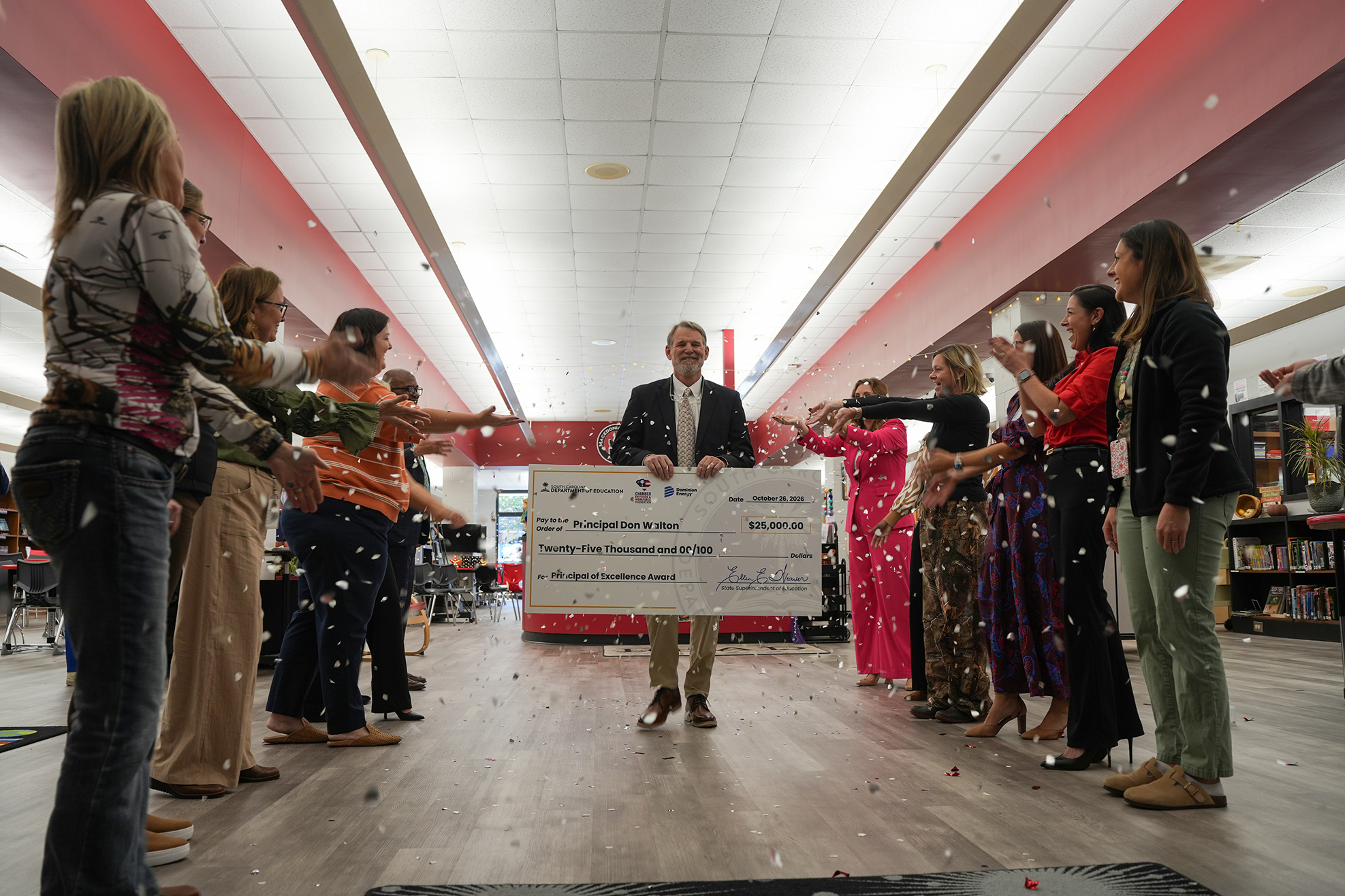 Adults in a school hallway stand around a man holding a large check. Confetti falls in the air.