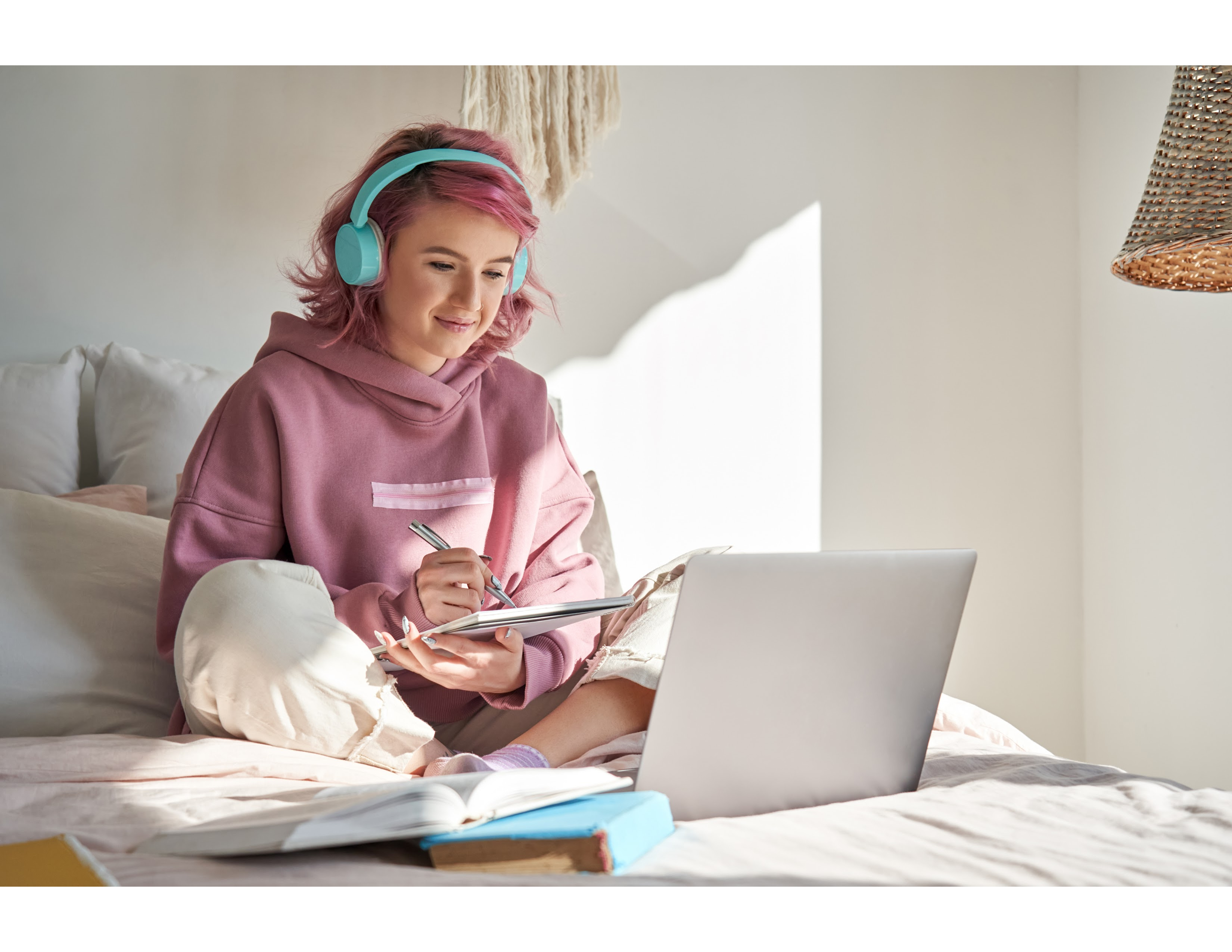 female student sitting on a bed with a laptop, notepad, and headphones