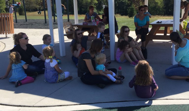 students sit under a pavillion in the shade and listen to a teacher read