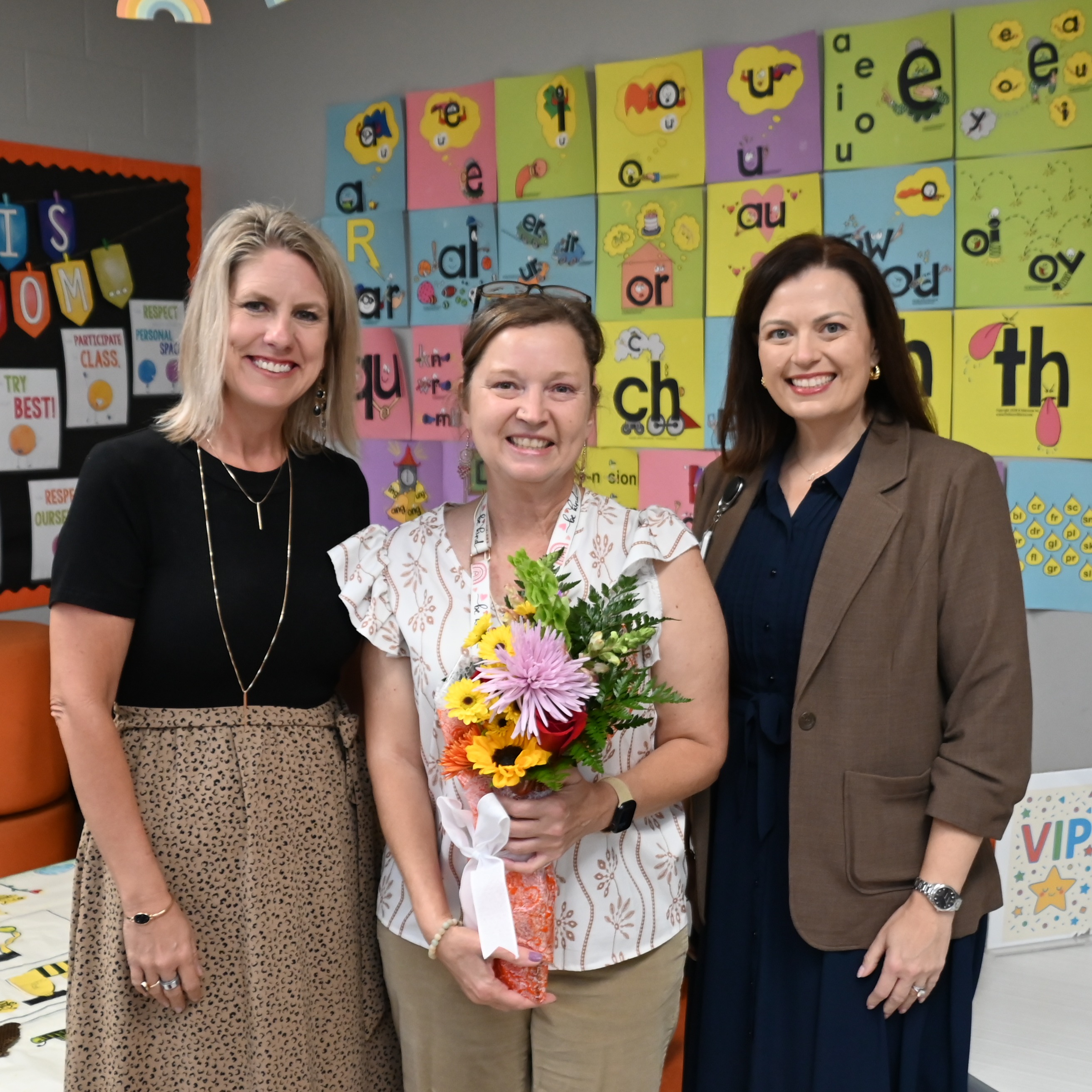 Three women stand in a room with a colorful alphabet wall in the background. The middle woman holds flowers.