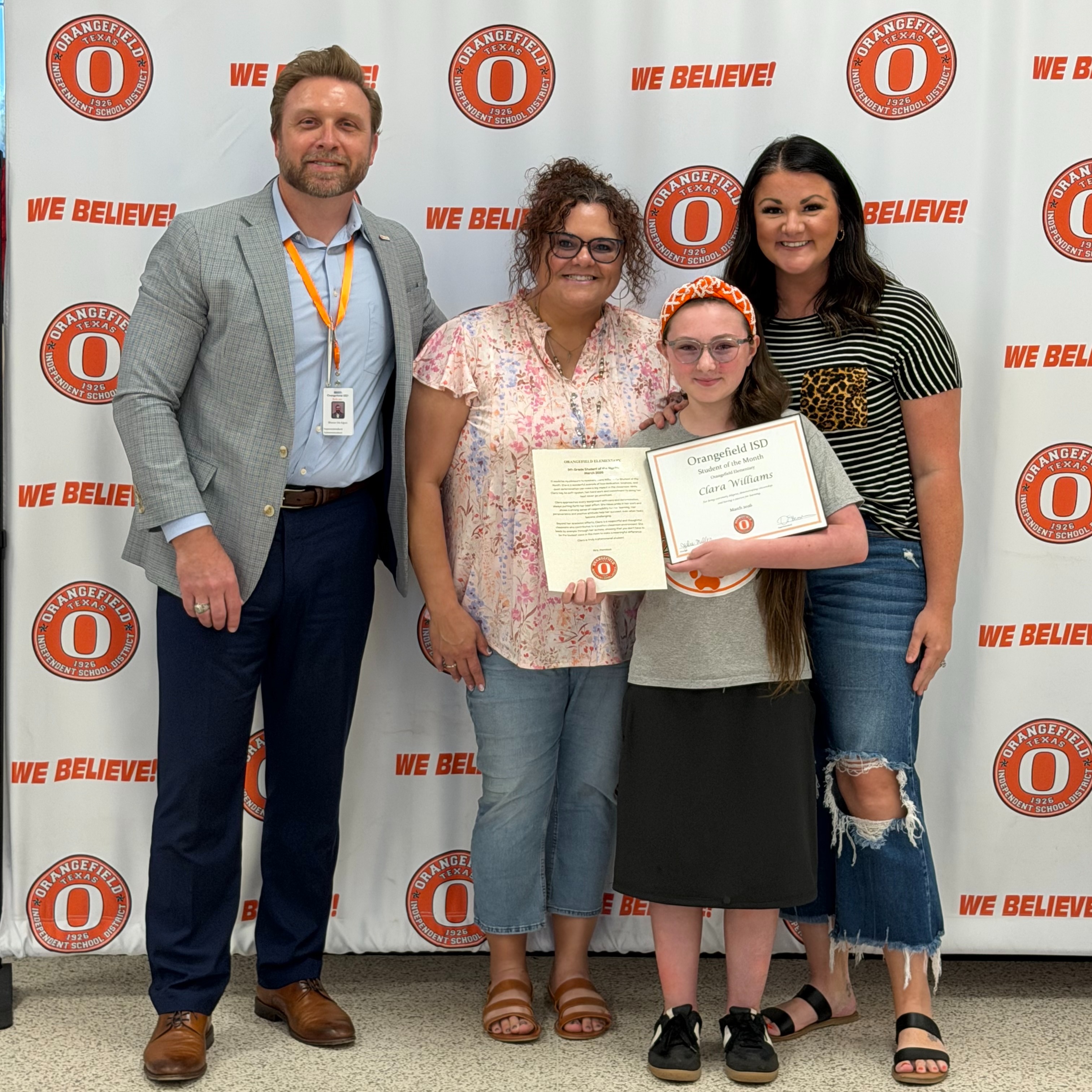 Five people stand in front of a white banner with "We Believe!" and an orange circle. A young girl holds a certificate.