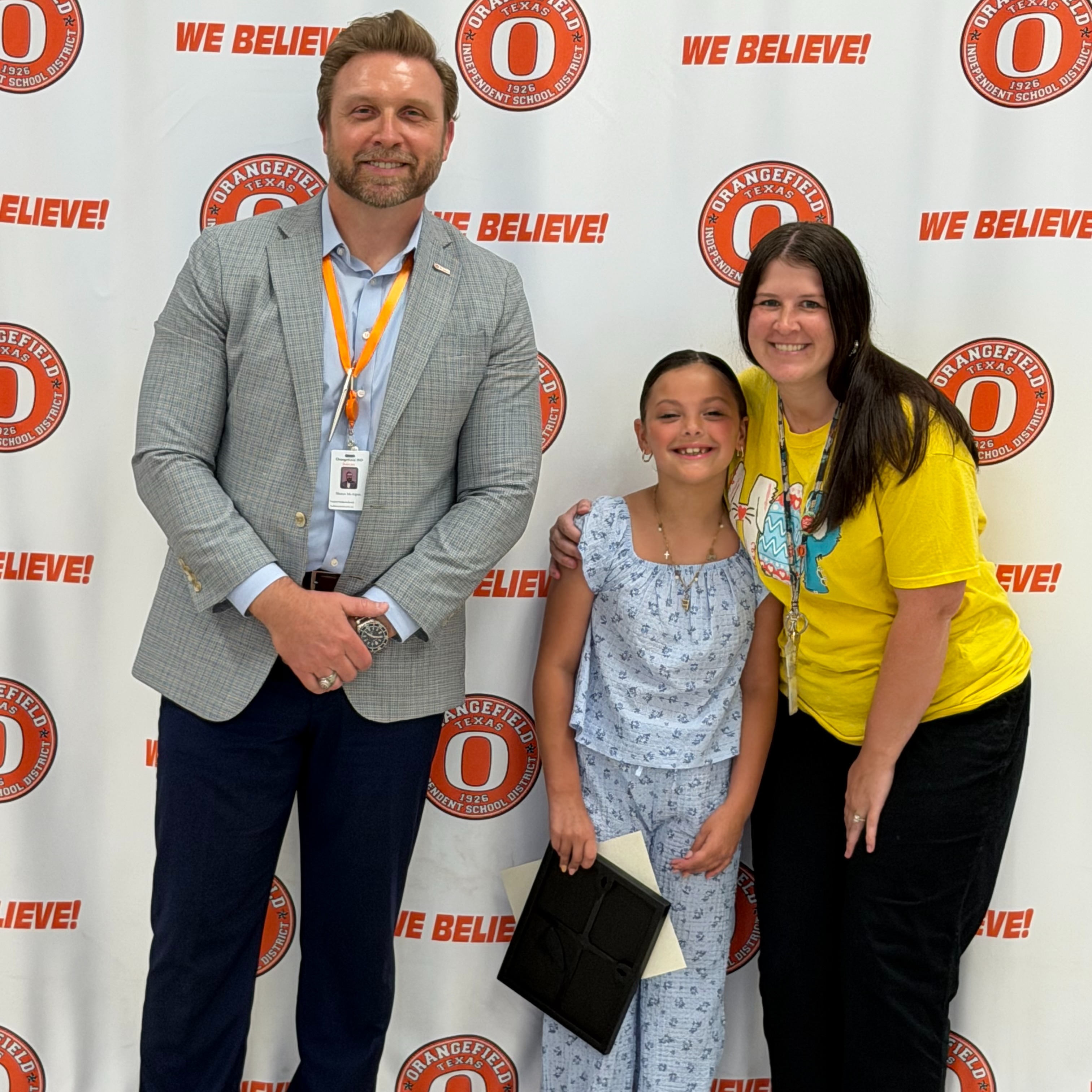 Three people standing with a banner behind them. A man in a gray suit, a young girl in pajamas, and a woman in a yellow shirt.