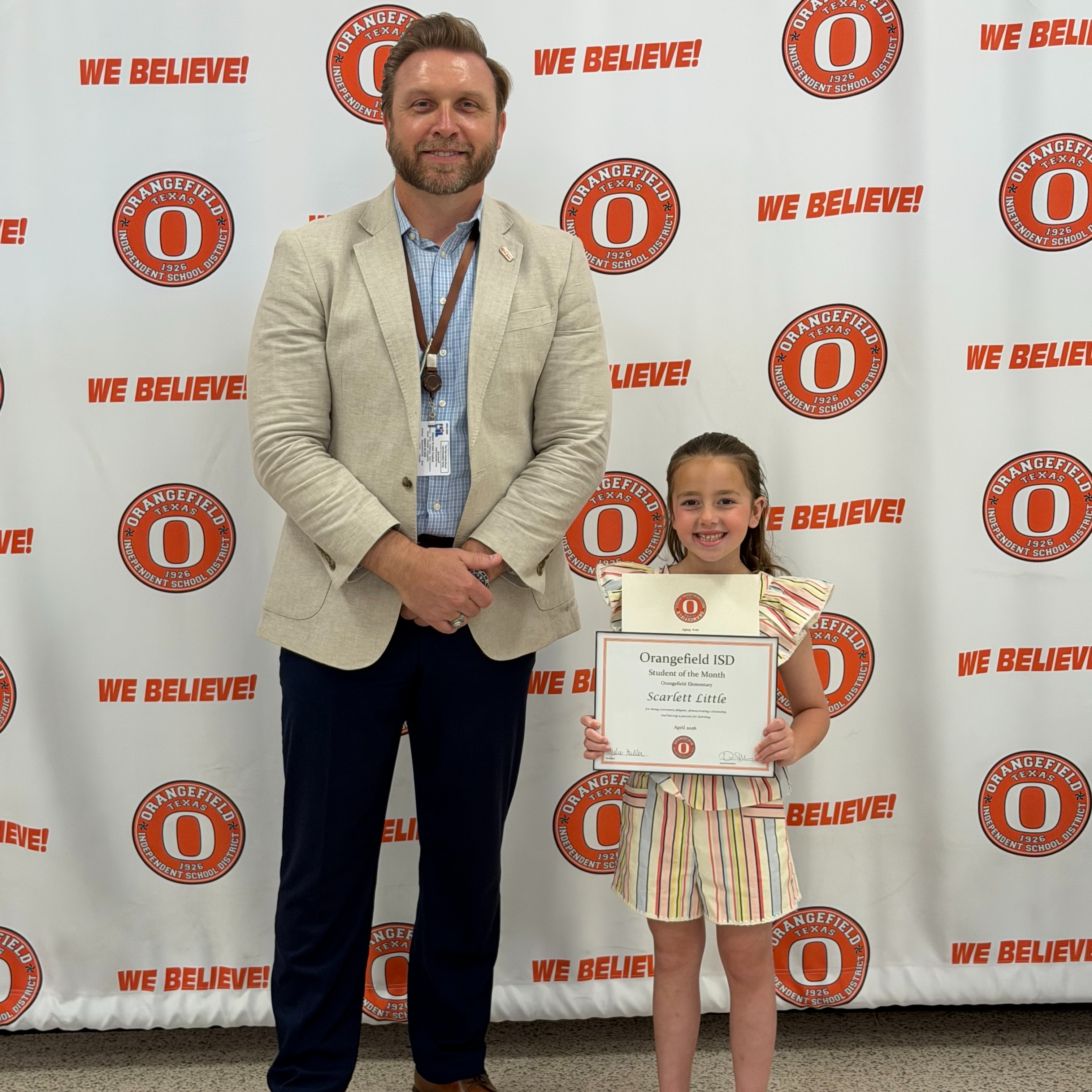 A man in a tan blazer and a young girl in a striped outfit hold a certificate before a wall with orange logos and the slogan "We Believe!"