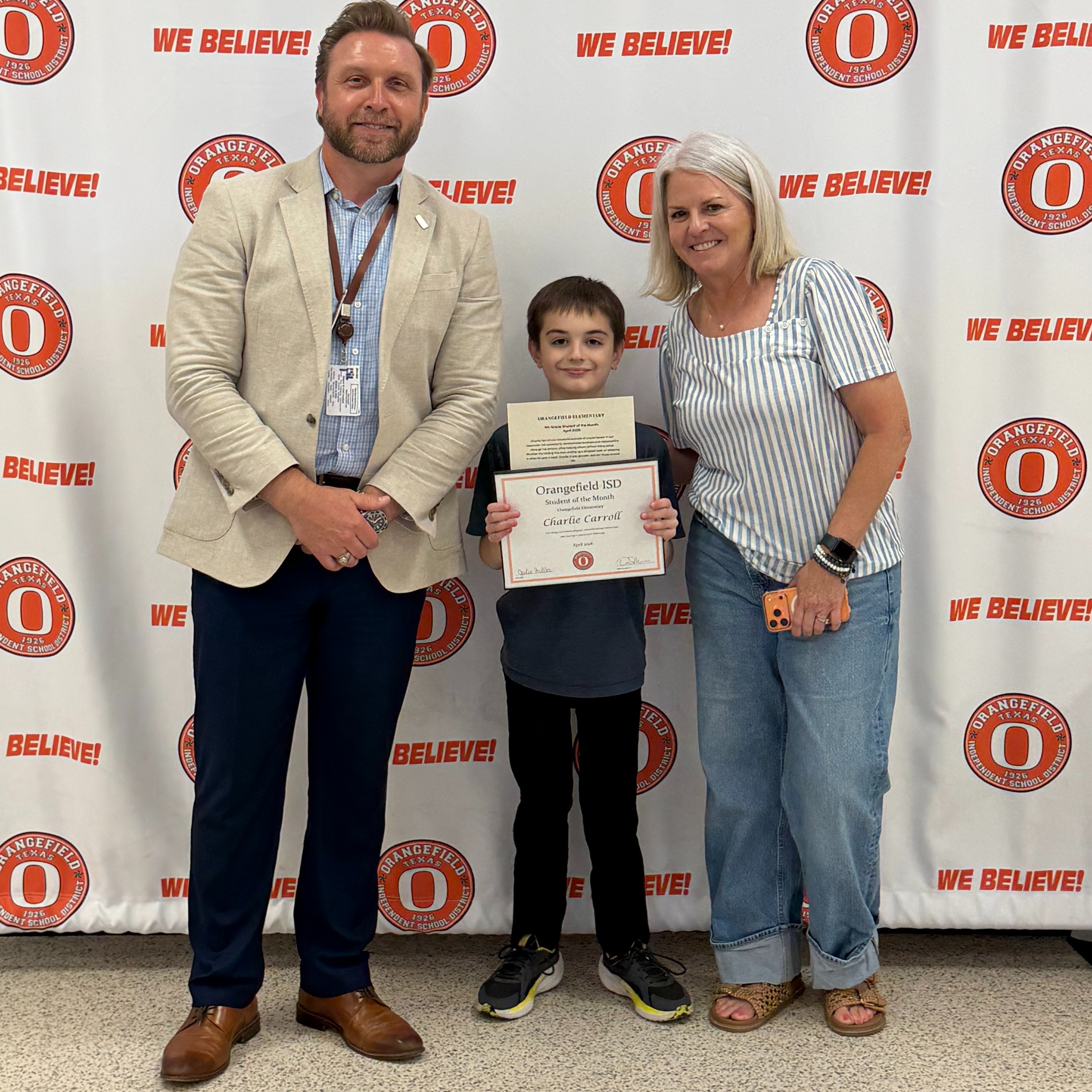 Three people stand together, one holding a certificate. They stand in front of a wall with large red letters.
