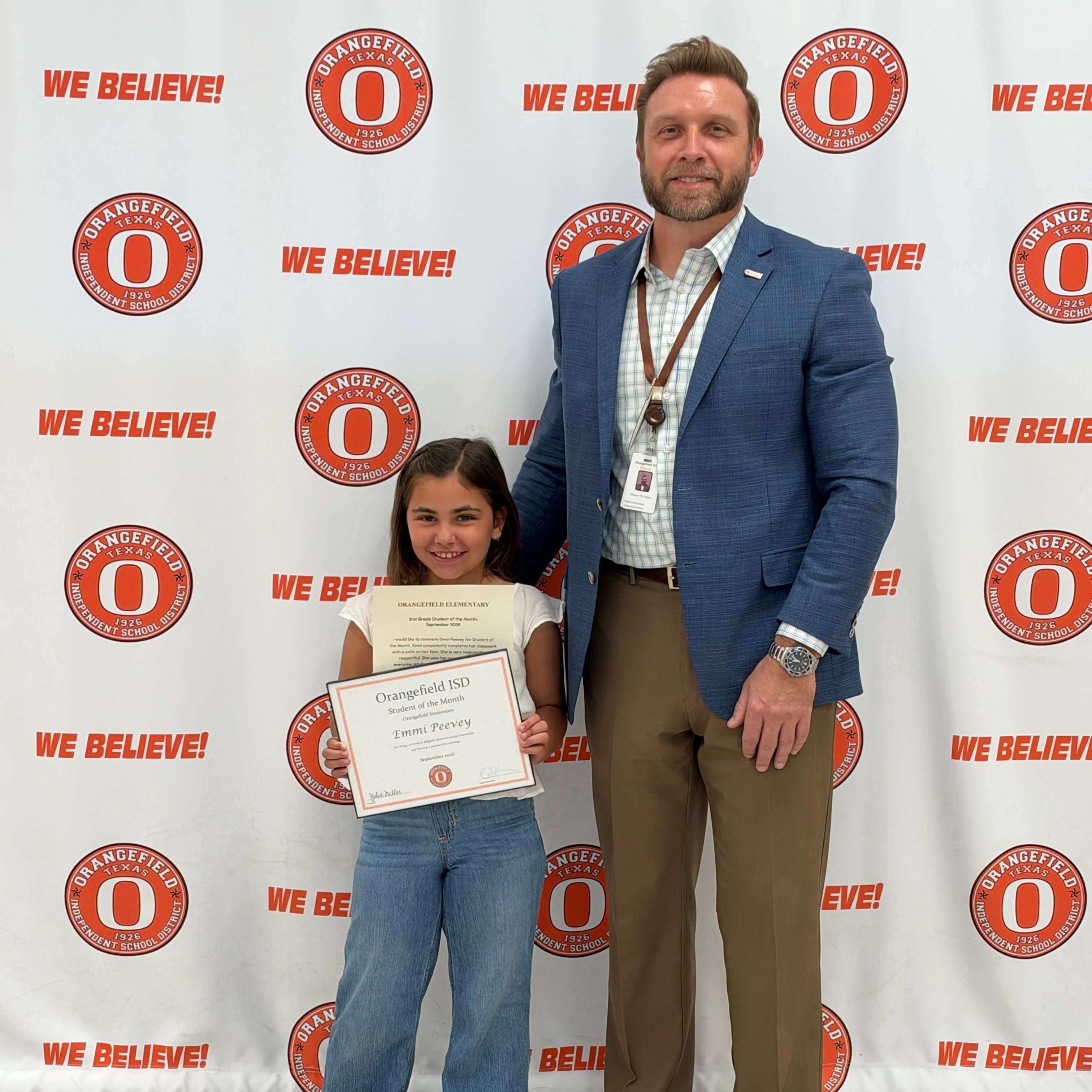 A young girl holding a certificate stands next to an adult man, both posing in front of a white backdrop with red logos.