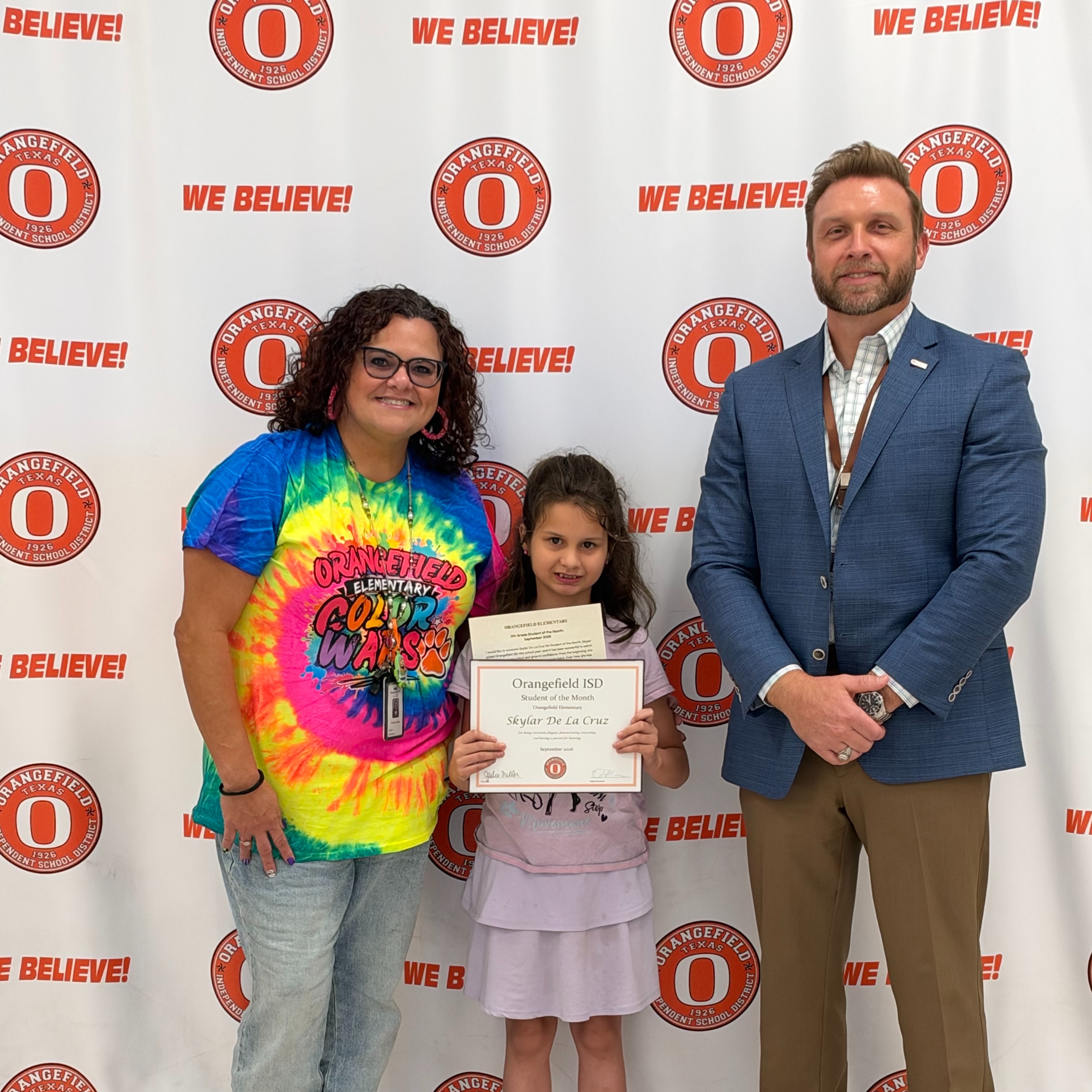 Two adults and a child stand in front of a wall with red text. The child holds a certificate.