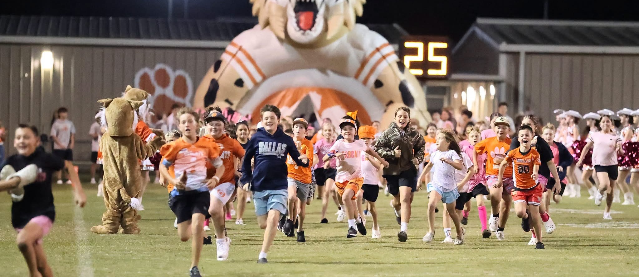 Children wearing orange shirts run on a field at night, with a large tiger mascot behind them.