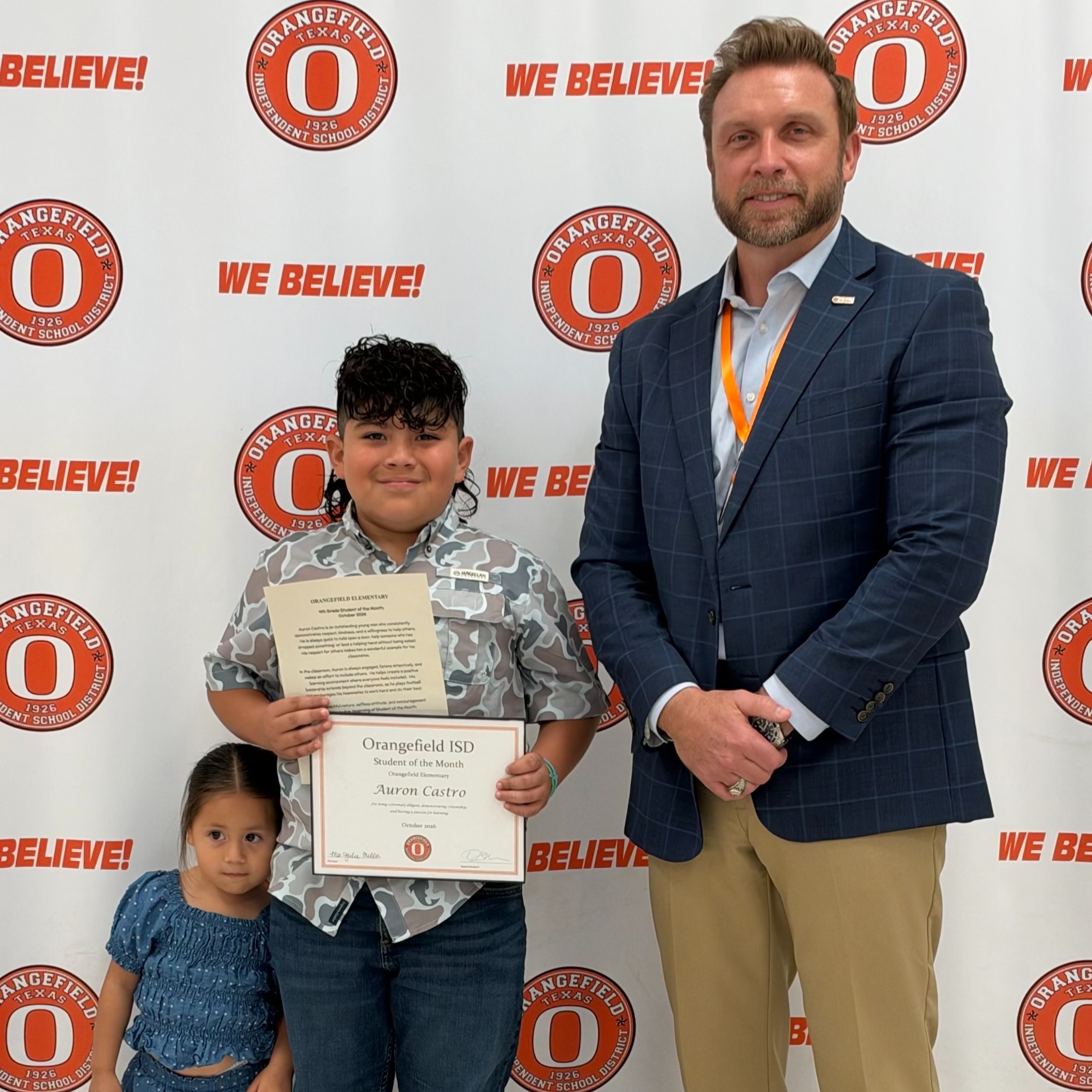 A boy holding a framed certificate stands between a man and a young girl. A white banner with "We Believe" printed on it is in the background.