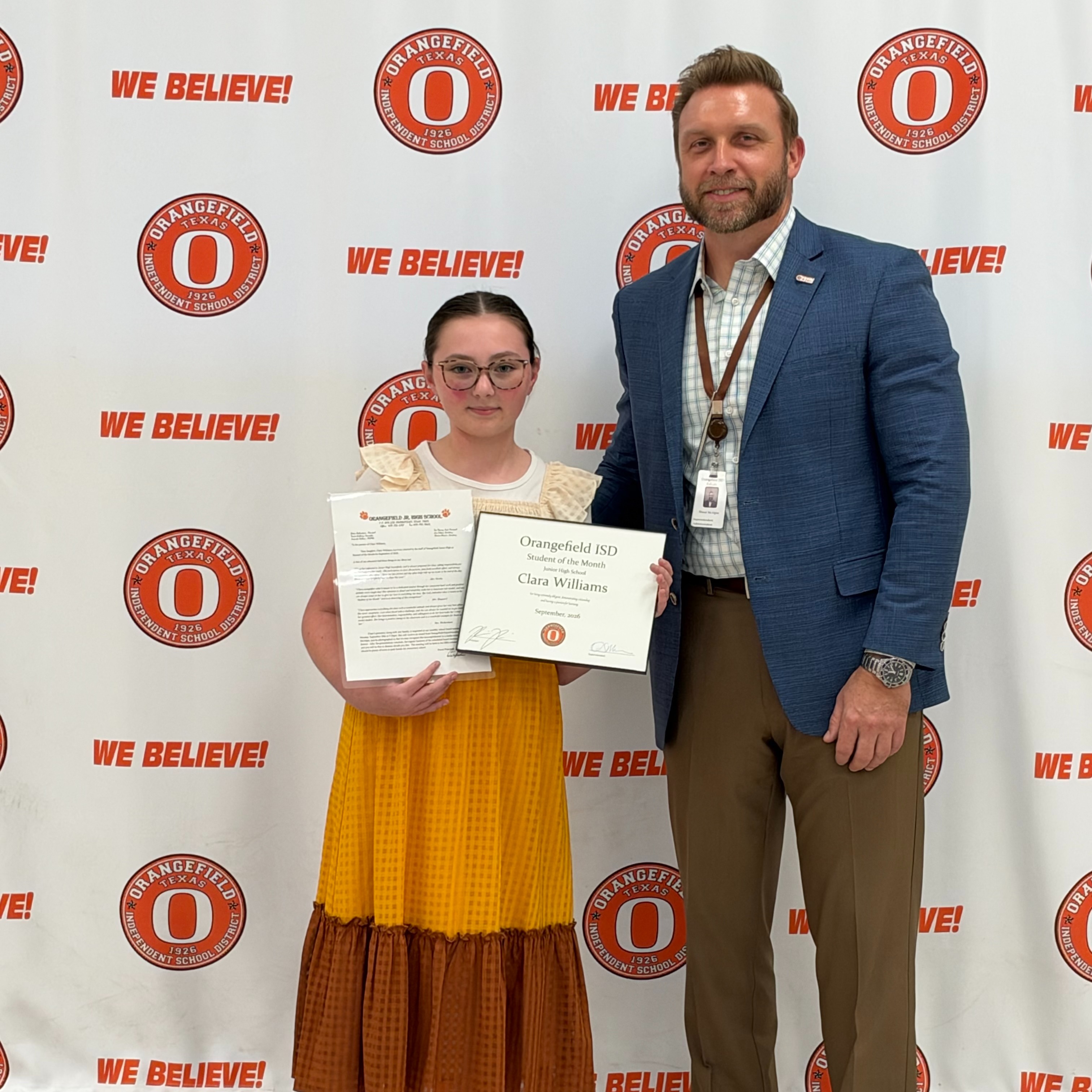 A man and a girl stand next to each other. Both hold certificates. They are in front of a banner.
