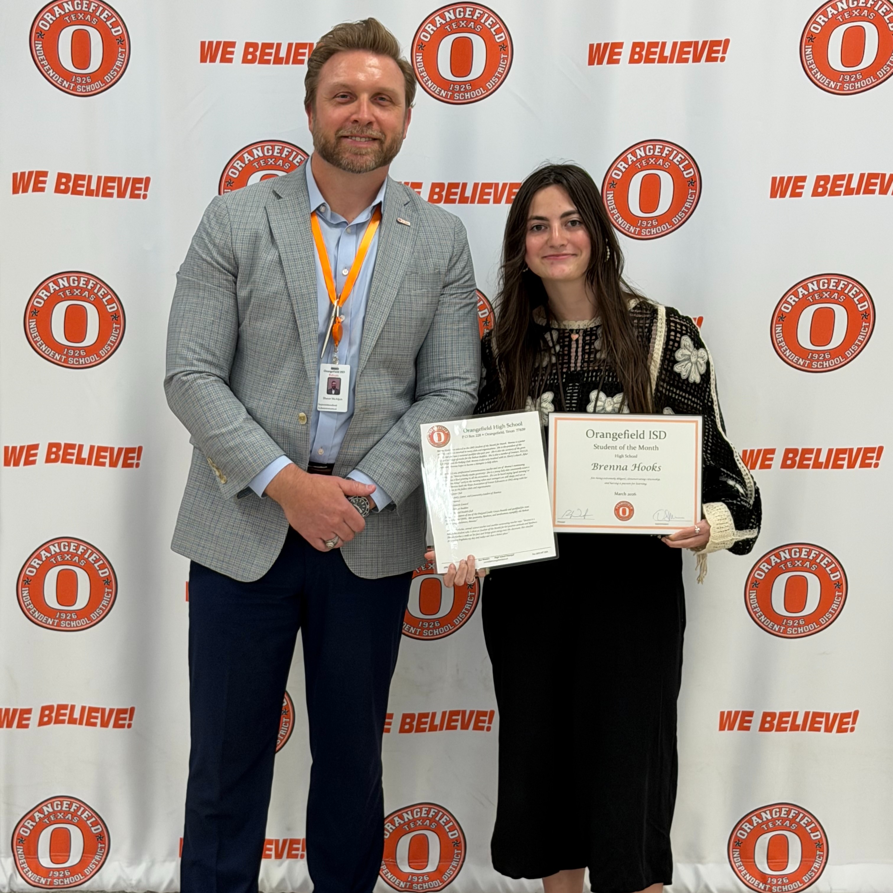 A man and a woman stand with certificates against a wall with orange circles and the text "We Believe."