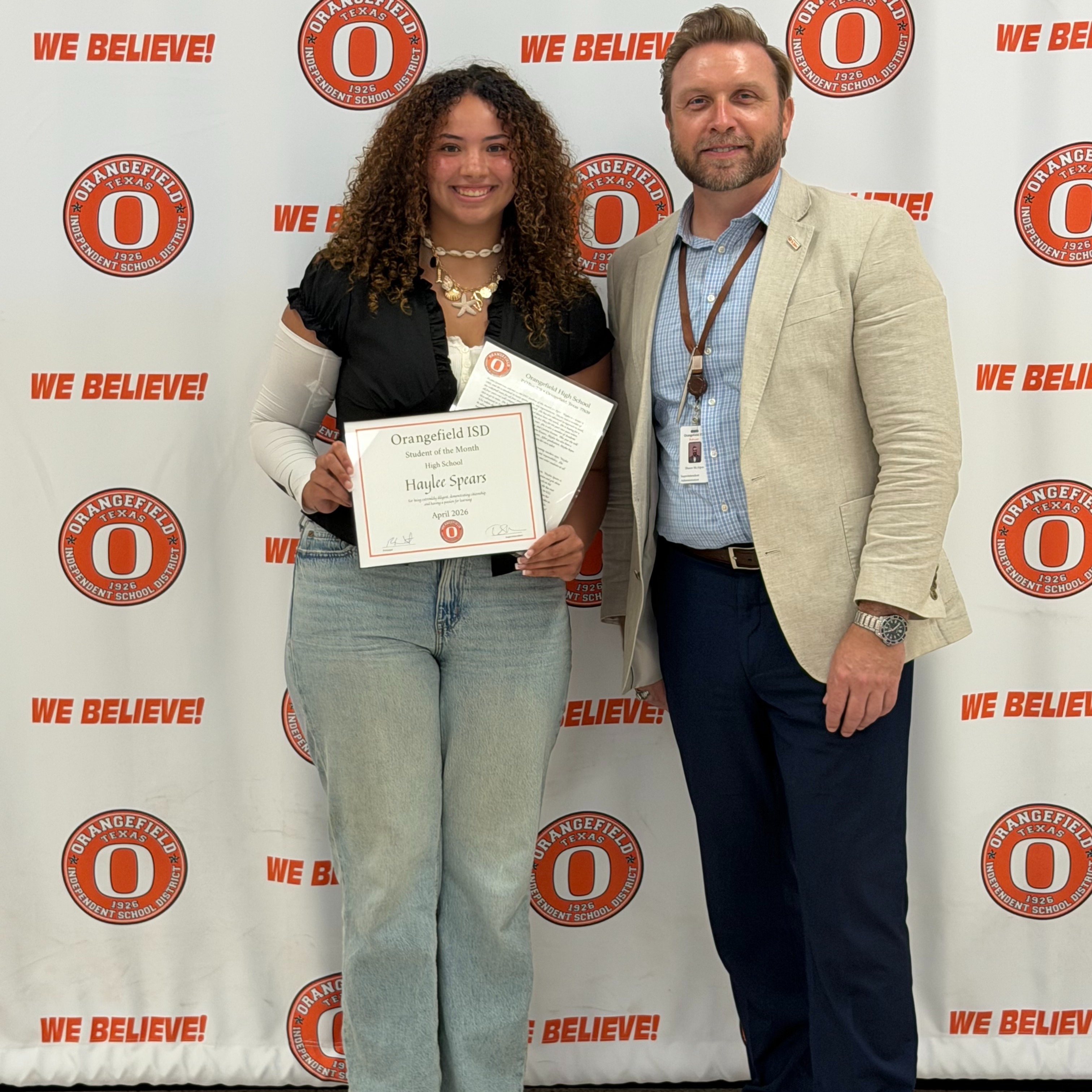 A woman holding a certificate stands next to a man in front of a backdrop. Both are smiling and looking at the camera.