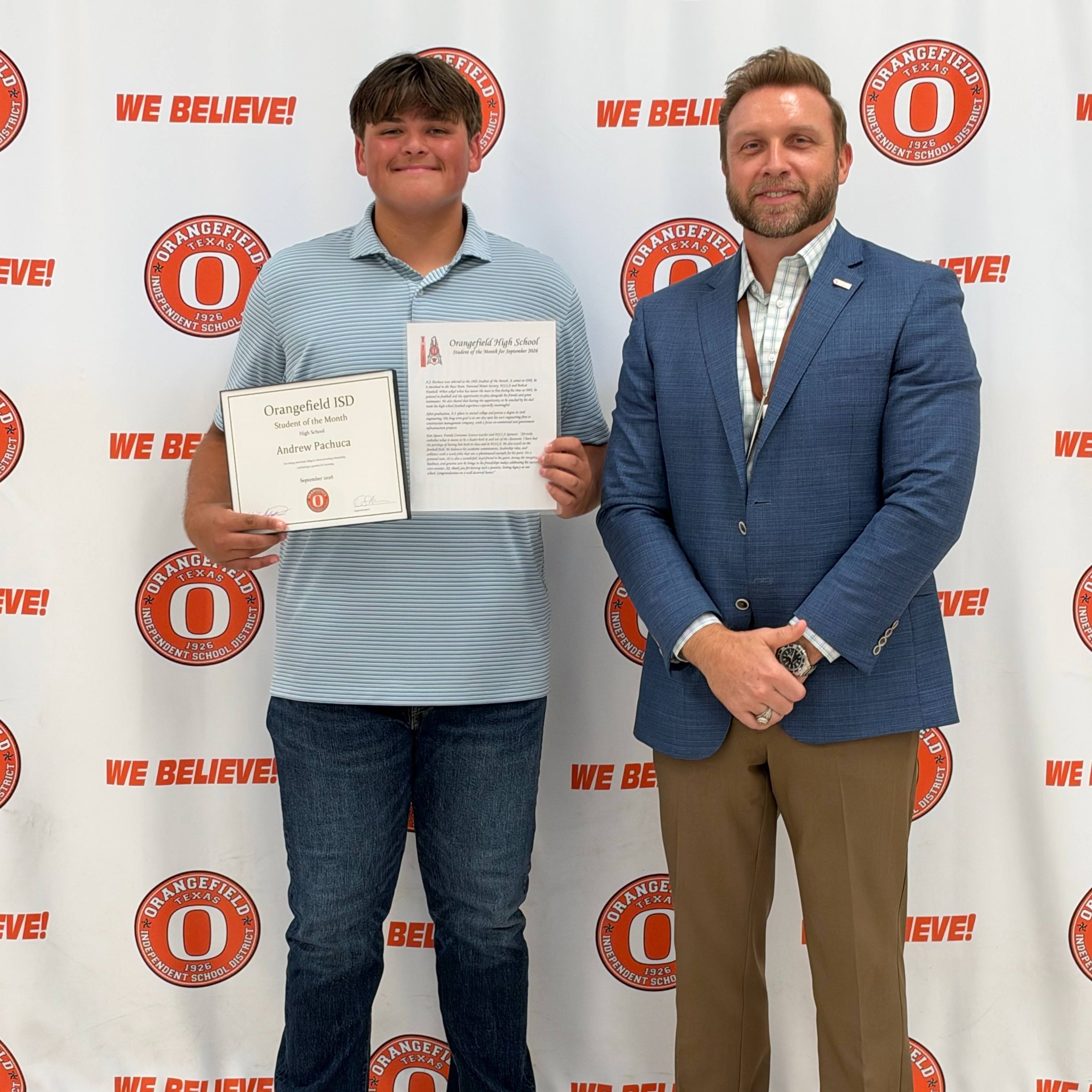 Two men stand before a wall with text and logos. One holds a certificate. The man on left wears a light blue shirt, blue jeans, and white sneakers. The man on right wears a blue suit, brown pants, and brown shoes.