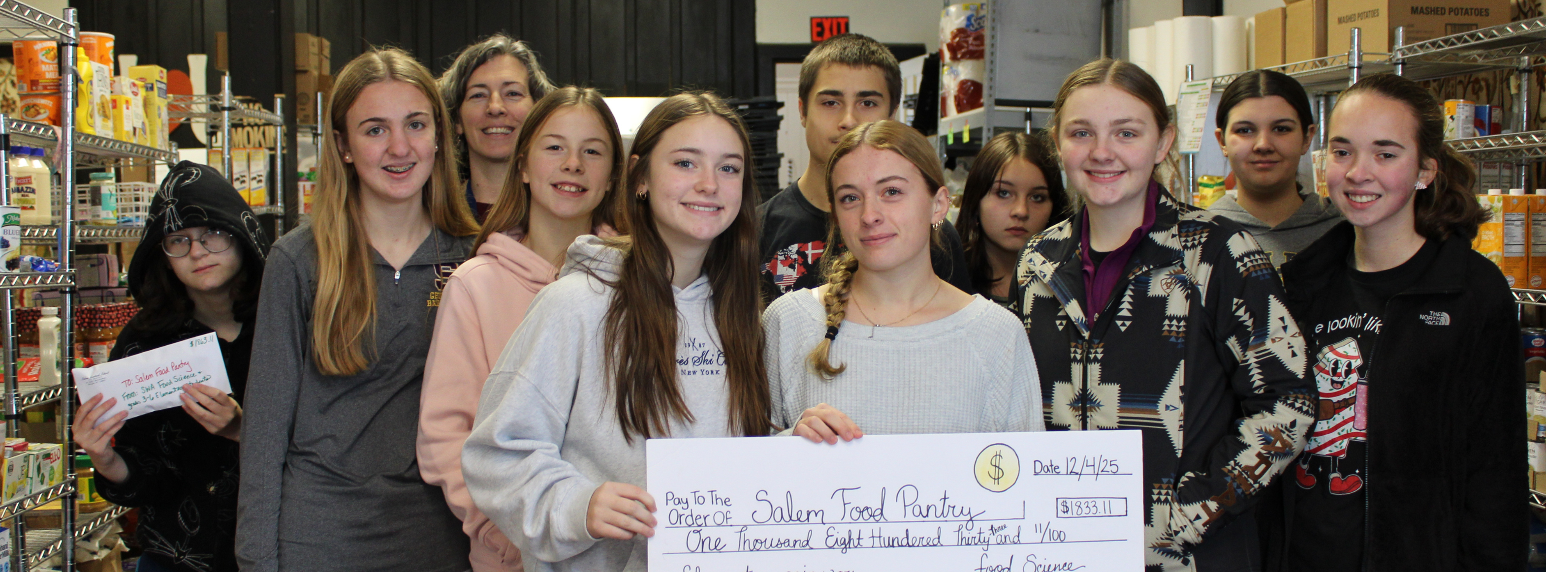 class of high school students stand in the food pantry and hold a giant check, representing the amount of money they donated