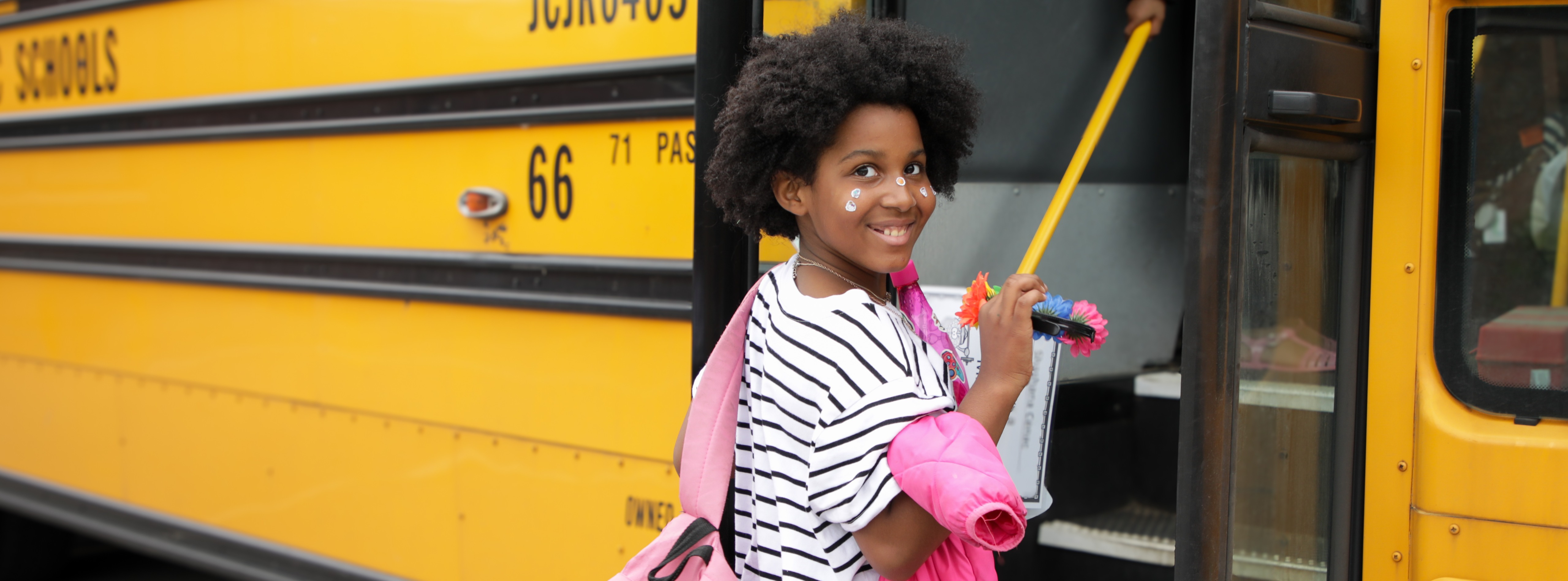 A child holding a pink coat and backpack smiles while boarding a yellow school bus.