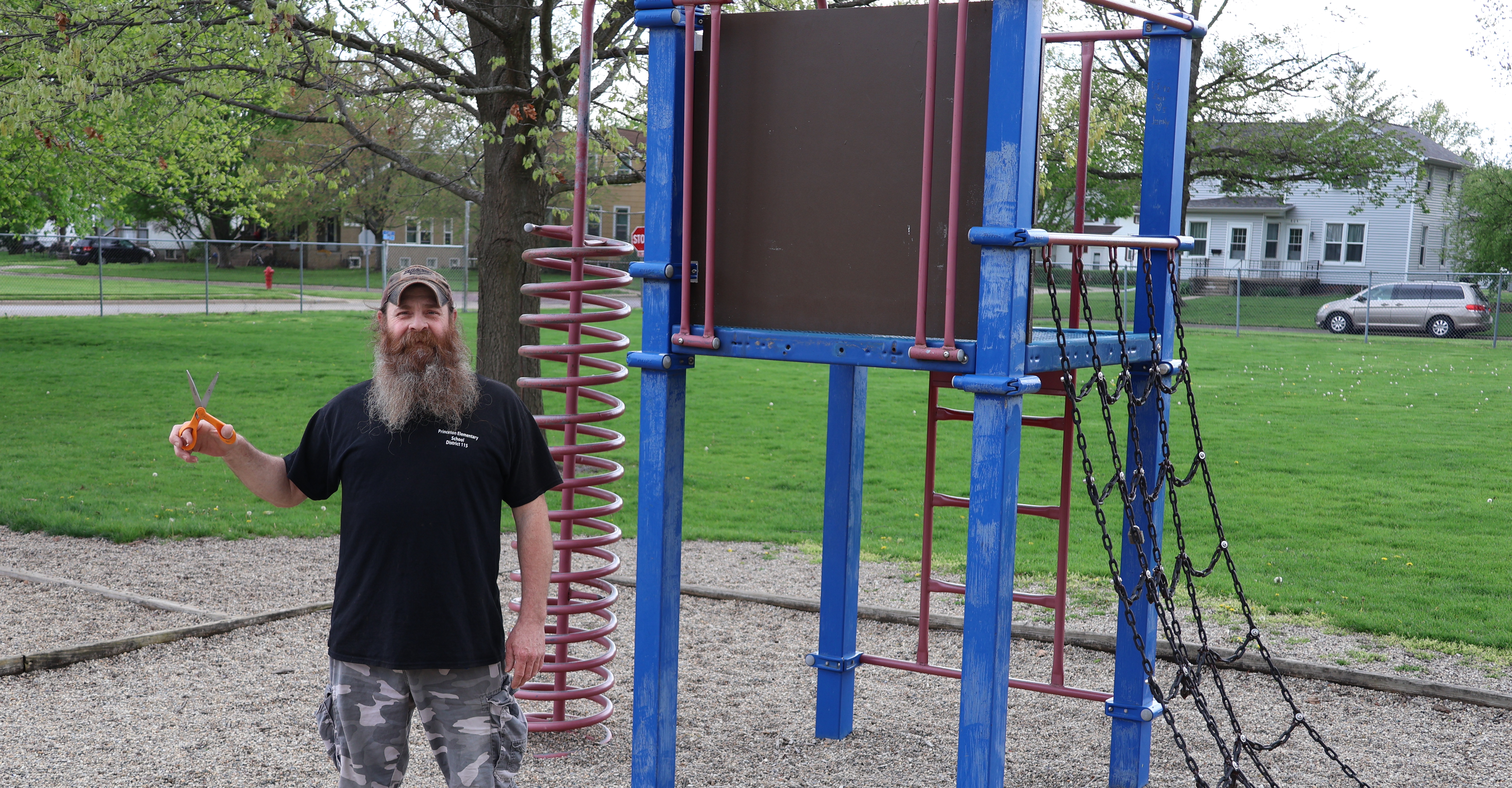 Custodian, Bob Espel, in front of old playground equipment with scissors in hand ready to cut his beard.