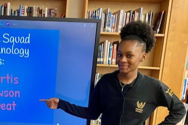 Iyana Sweat pointing at Interactive Board that Reads "Pheonix Tech Squad Student Technology Leaders: Piper Curtis Isaiah Lawson Iyana Sweat