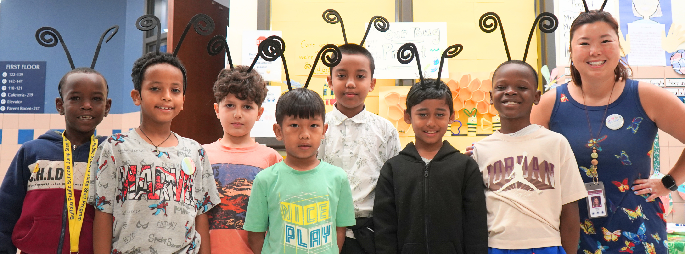Group photo of several students standing indoors with an adult in a school hallway or common area. The students are wearing headband antennae as part of a themed activity or display. Posters and classroom signage are visible in the background, suggesting a school event or project showcase.