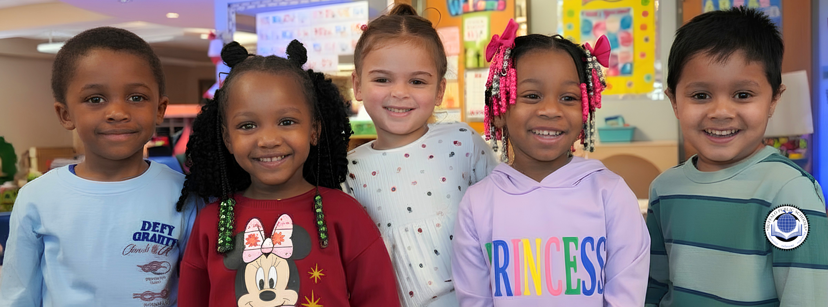Group photo of several students standing indoors with an adult in a school hallway or common area. The students are wearing headband antennae as part of a themed activity or display. Posters and classroom signage are visible in the background, suggesting a school event or project showcase.