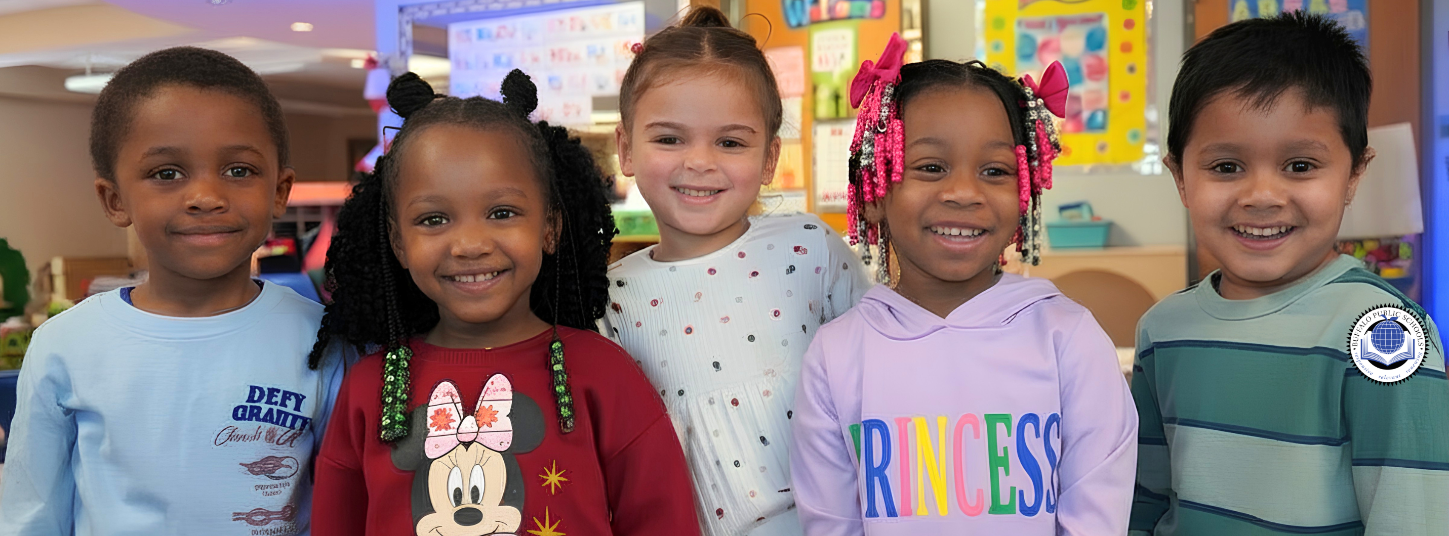 Group photo of several students standing indoors with an adult in a school hallway or common area. The students are wearing headband antennae as part of a themed activity or display. Posters and classroom signage are visible in the background, suggesting a school event or project showcase.
